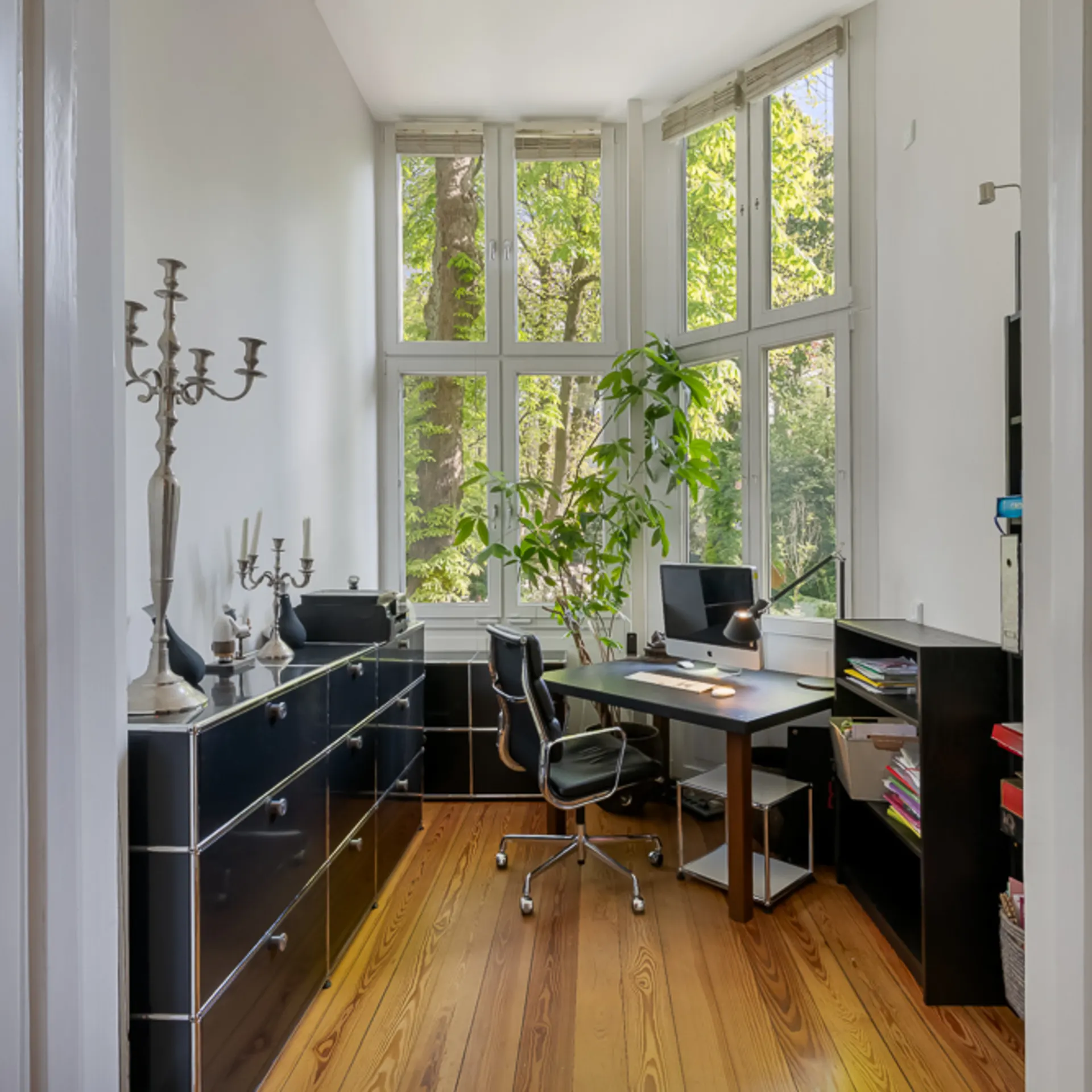 Helles Arbeitszimmer mit Schreibtisch, Computer, Regalen und großen Fenstern mit Blick ins Grüne.