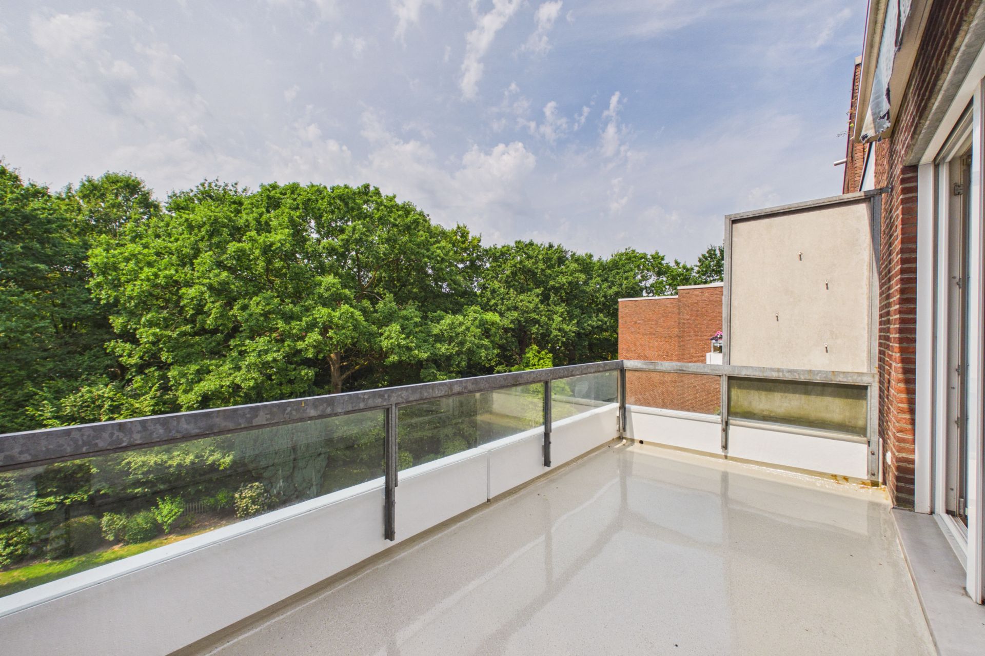 Geräumiger Balkon mit Glasgeländer, Blick auf grüne Bäume und angrenzende Gebäude.