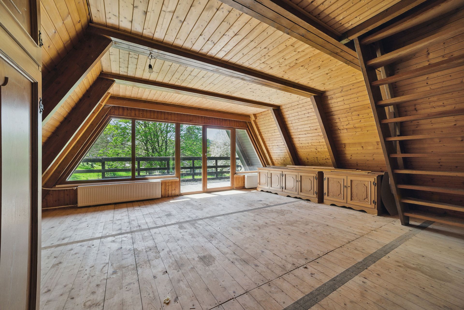 Großer Dachraum mit Holzwänden, Fenstern und Zugang zu einem Balkon, Holztreppe an der Seite.