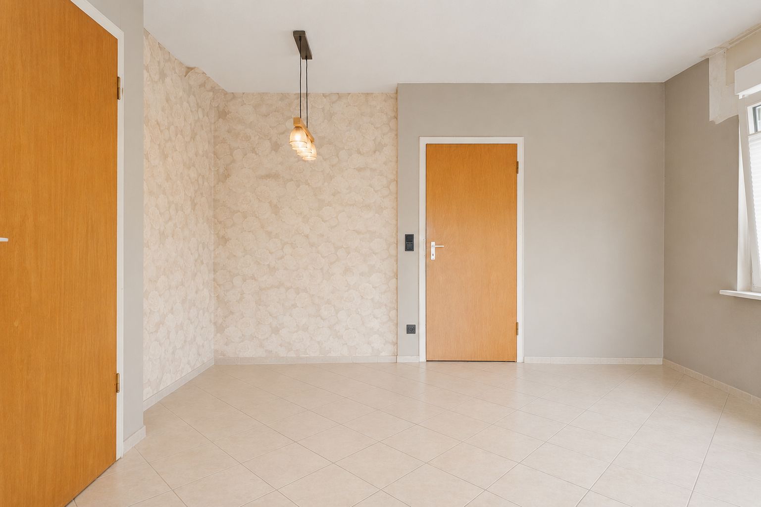 Heller, leerer Raum mit beigen Fliesen, Holztüren, Fenster und zwei Deckenlampen.