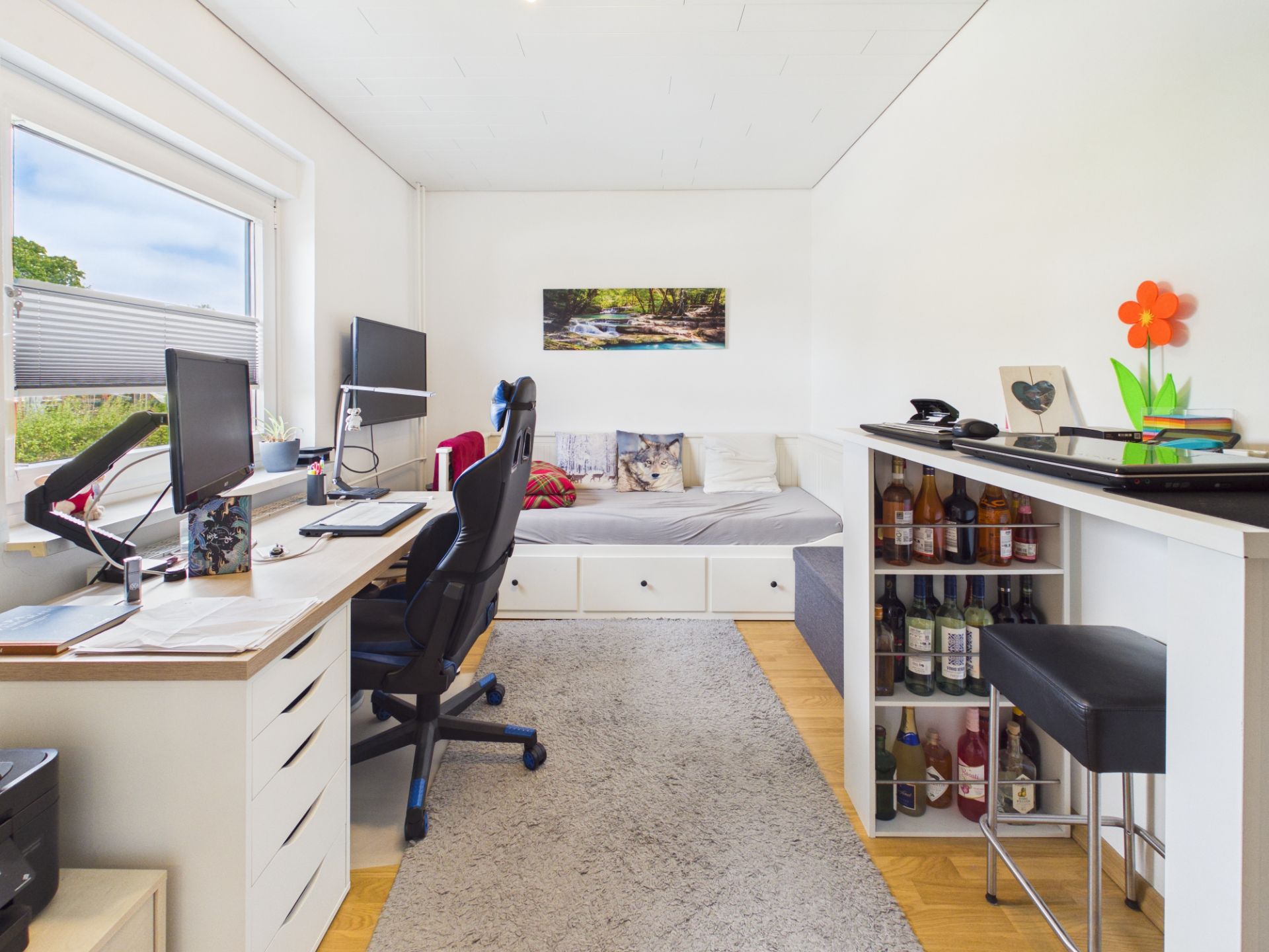 Helles Zimmer mit Schreibtisch, Bürostuhl, Tagesbett und Regal mit Flaschen.