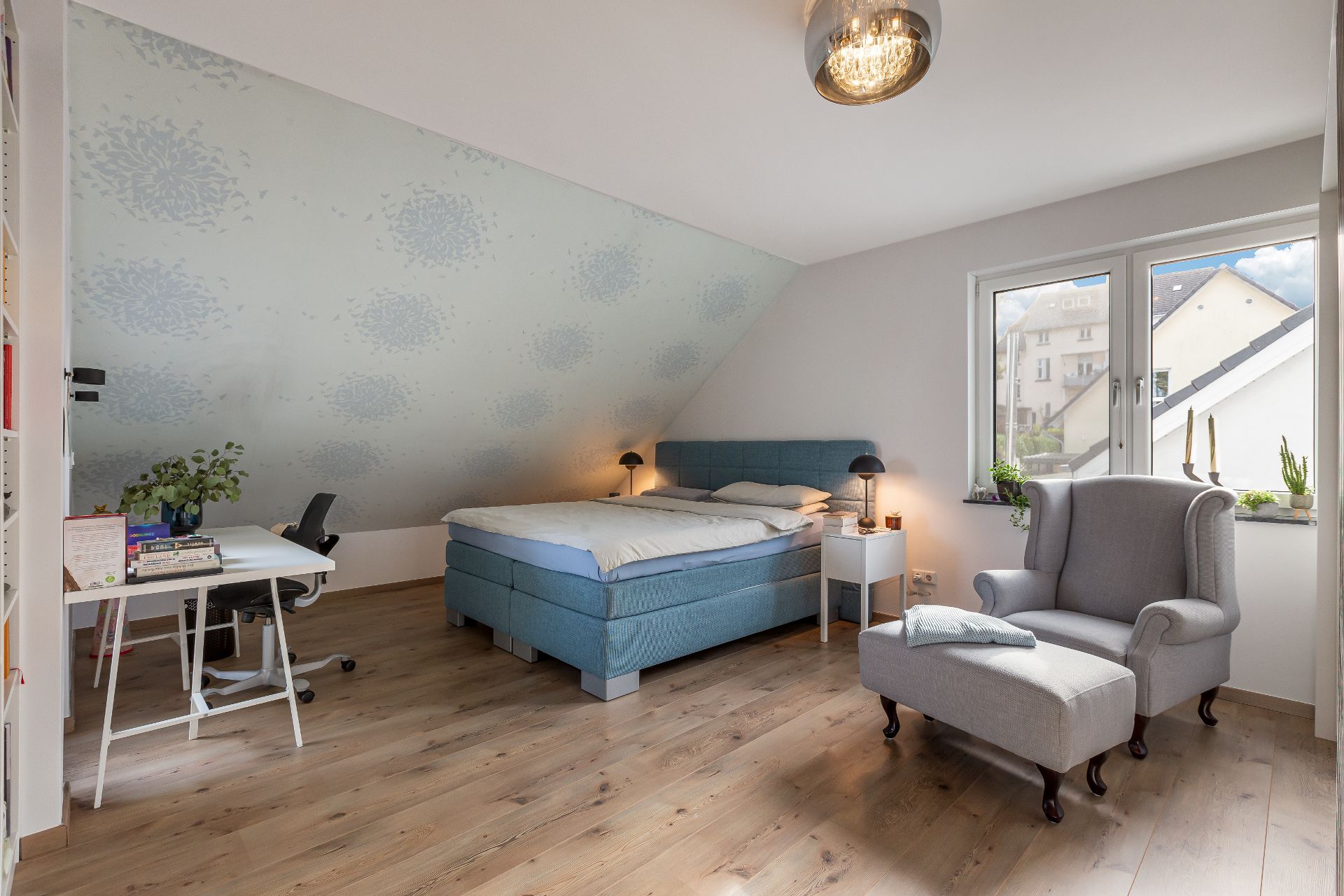 Schlafzimmer mit Doppelbett, Schreibtisch, Sessel und zwei Fenstern, Holzfußboden.