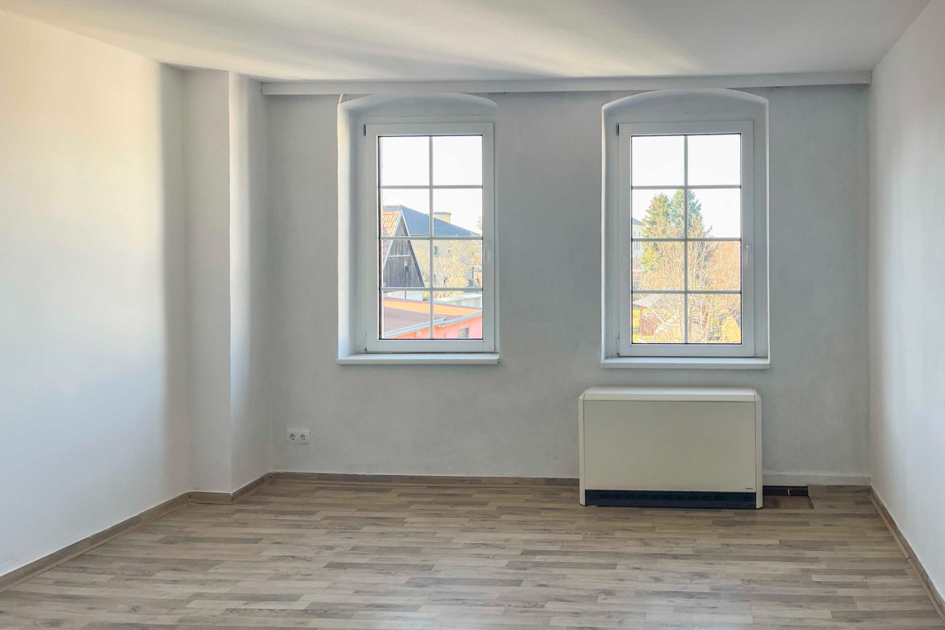 Heller Raum mit zwei Fenstern, weiß gestrichenen Wänden und hellem Holzfußboden.