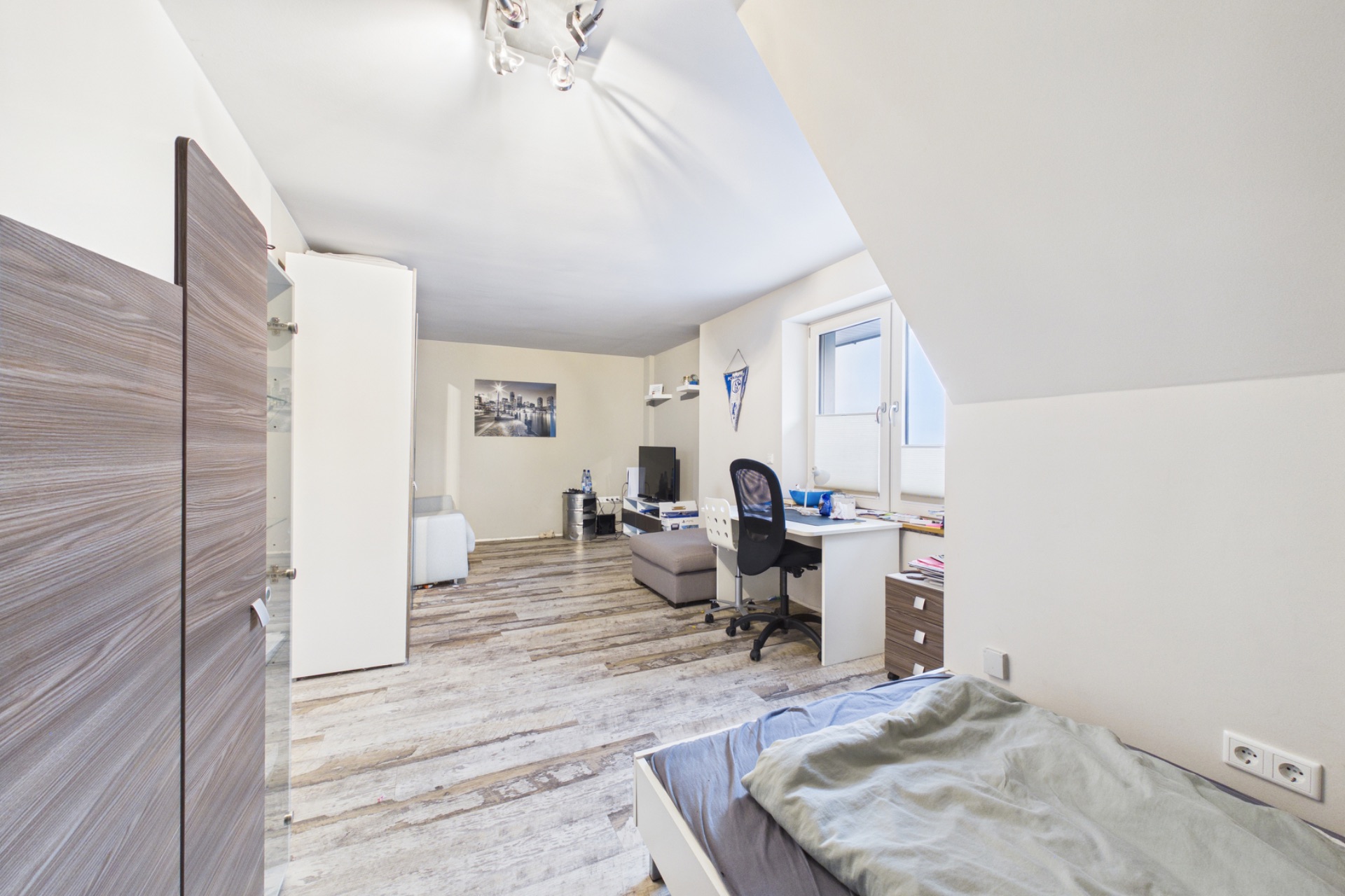 Helles Schlafzimmer mit Bett, Schreibtisch, Stuhl und Sessel auf Holzboden.