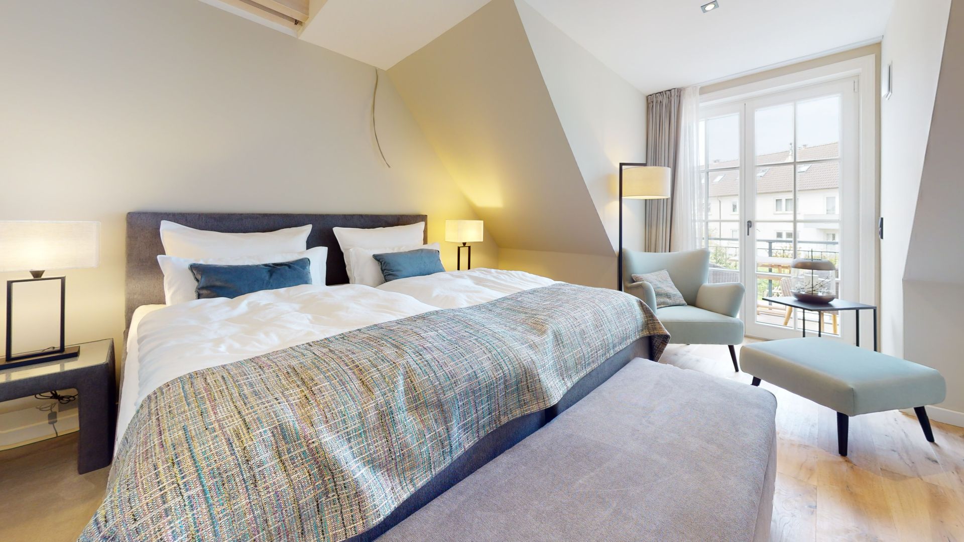 Helles Schlafzimmer mit Doppelbett, Sessel, Hocker und großem Fenster mit Blick nach draußen.