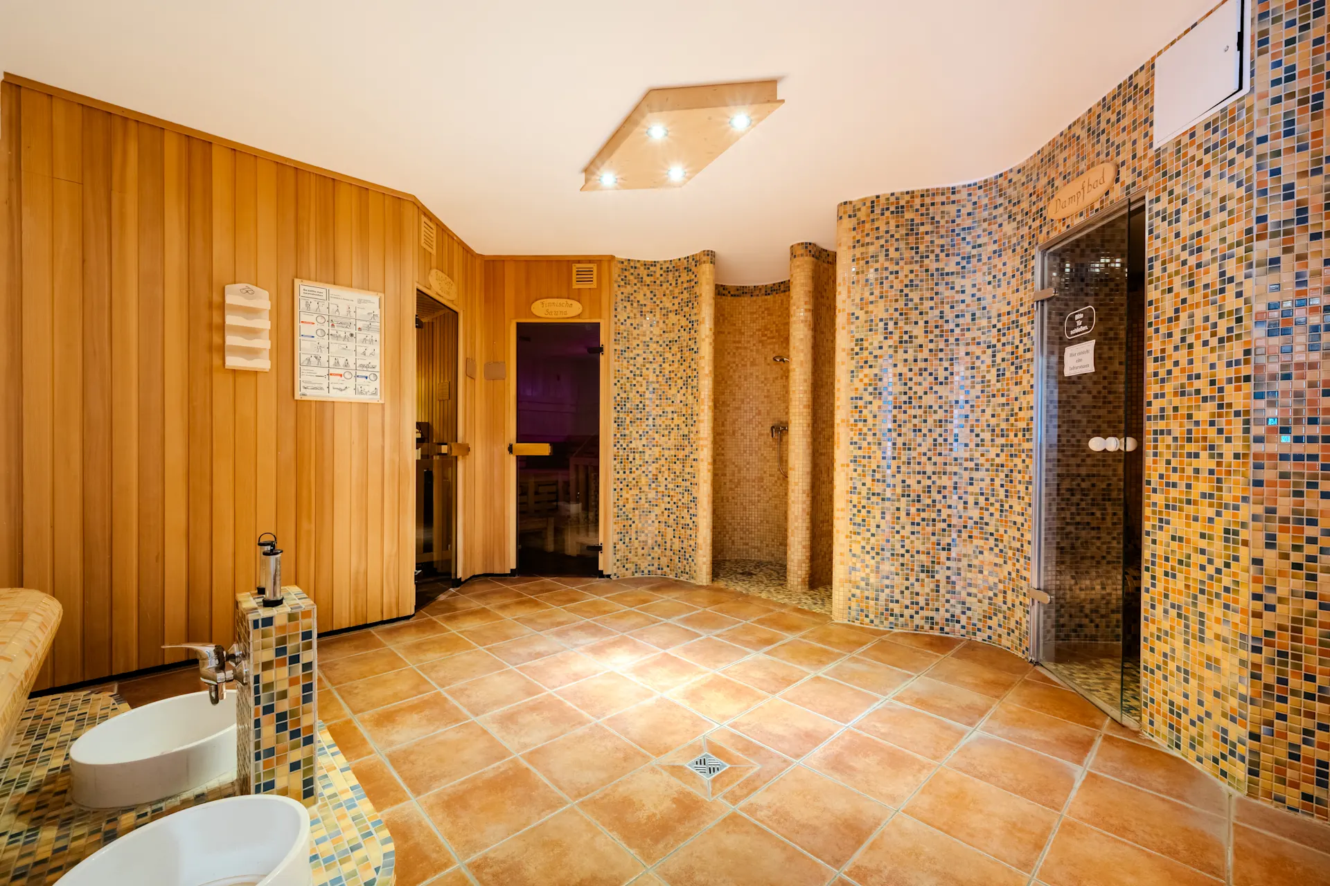 Wellnessbereich mit Holzwänden, gefliesten Duschen und einer Glastür zu einer Sauna.