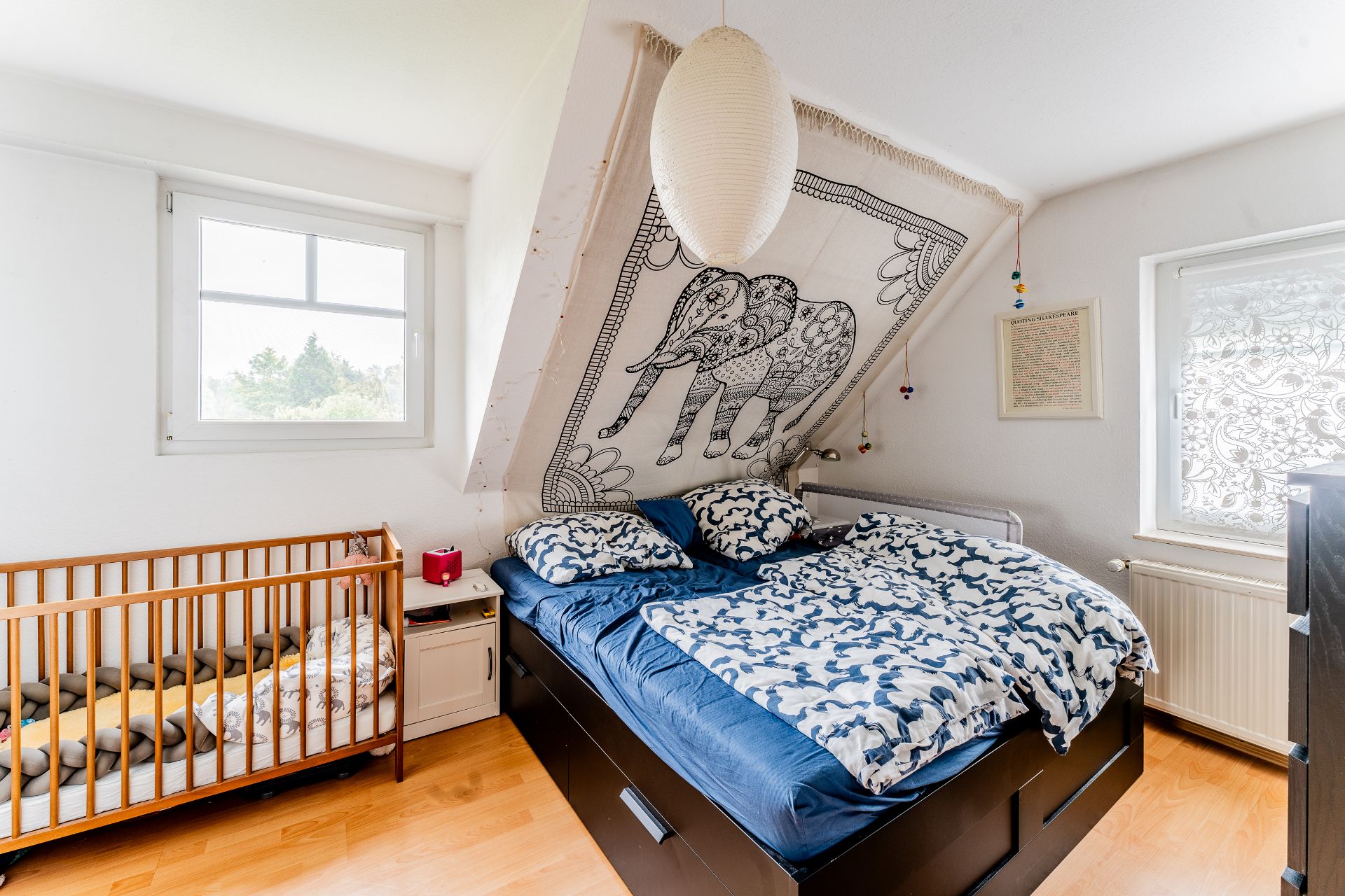 Schlafzimmer mit Doppelbett, Babybett und Elefanten-Wandteppich unter einer Dachschräge.