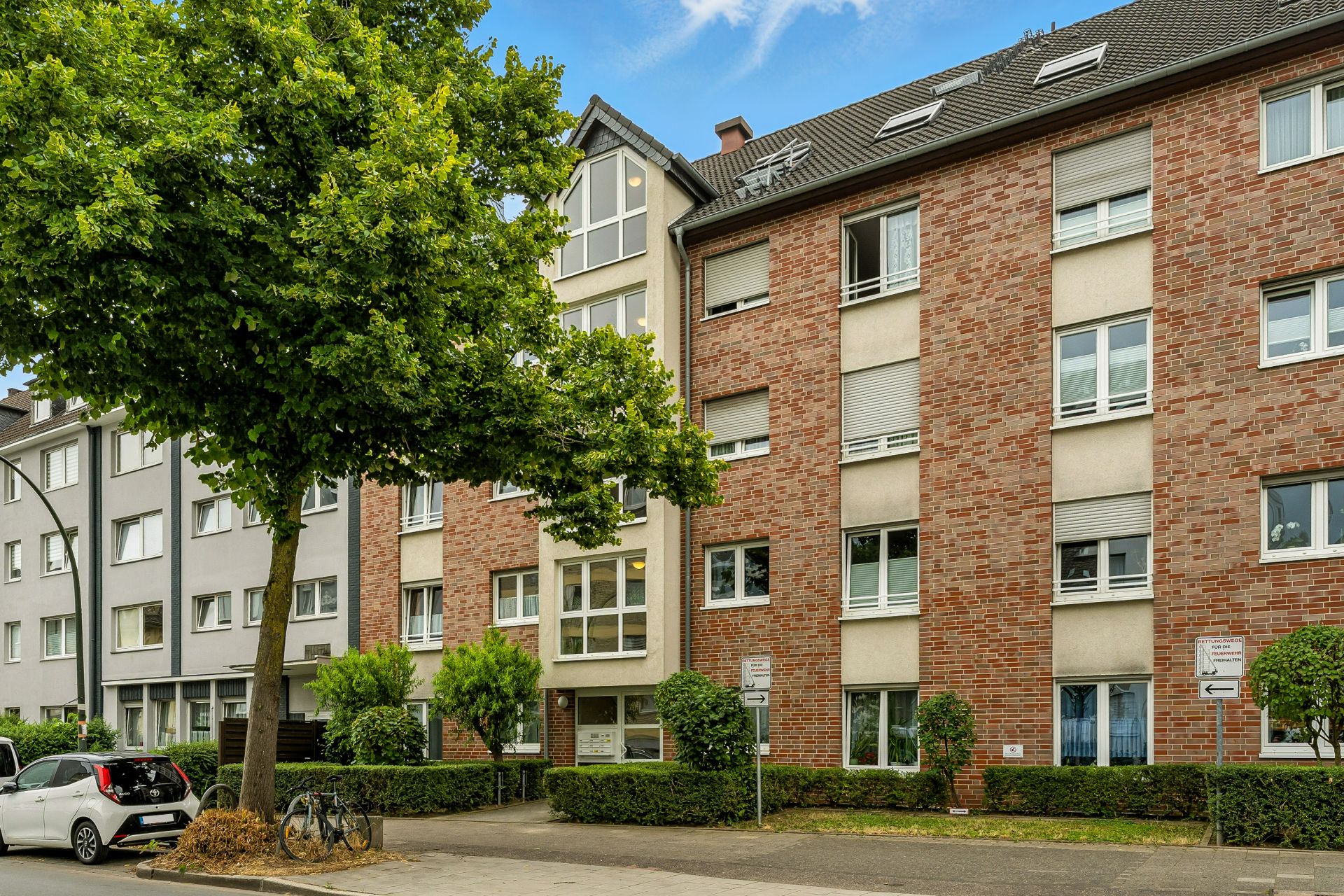 Mehrfamilienhaus mit roter Fassade, Bäumen und geparkten Autos auf der Straße.