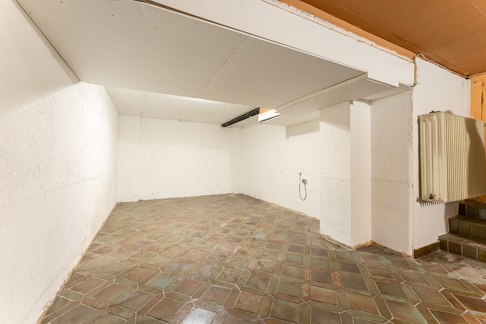 Leerer Kellerraum mit gefliestem Boden, weißen Wänden und einer Treppe mit Heizkörper.