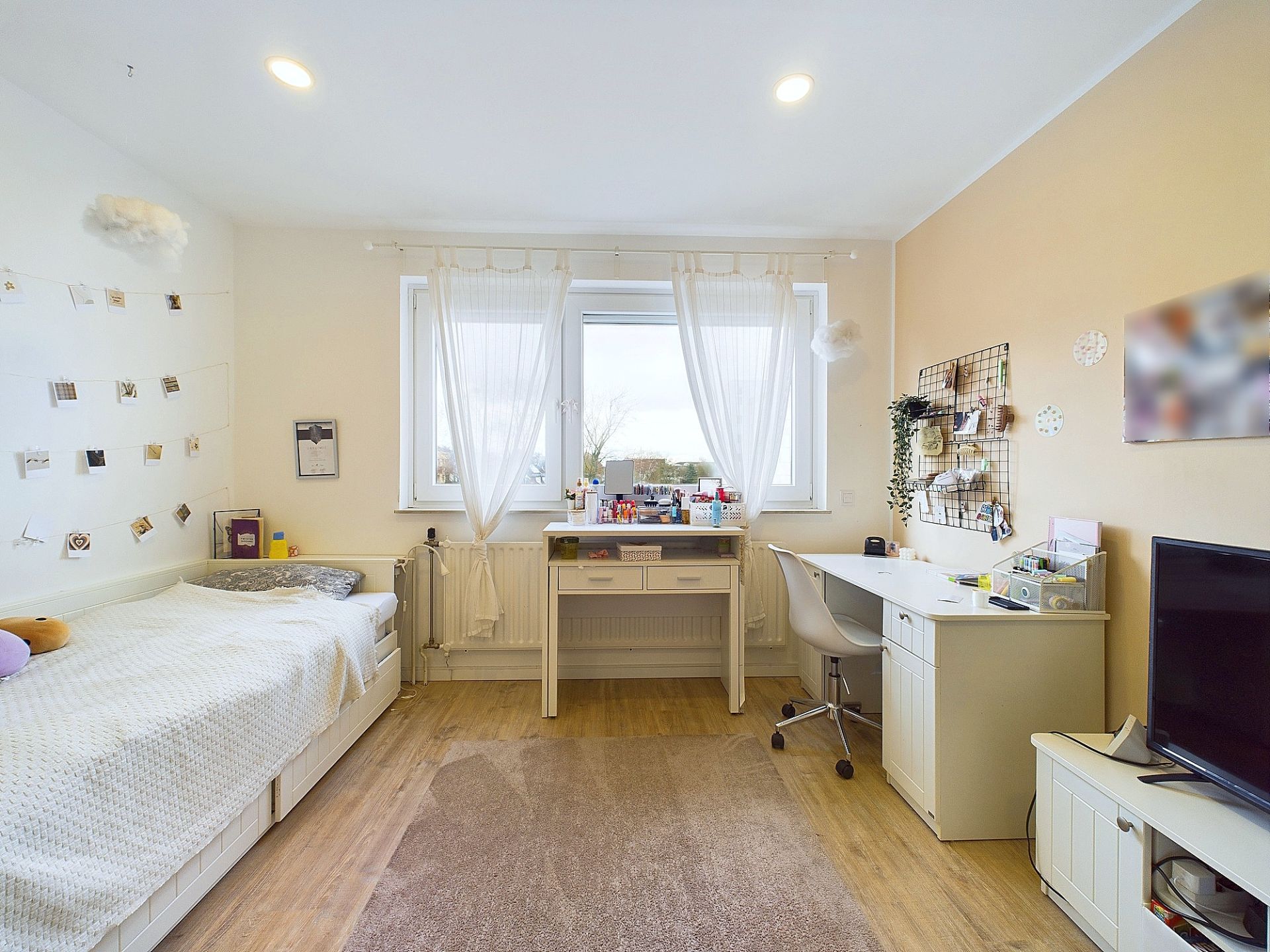 Helles Schlafzimmer mit Bett, Schreibtischen, Stuhl, Fenster mit Gardinen und Fernseher.