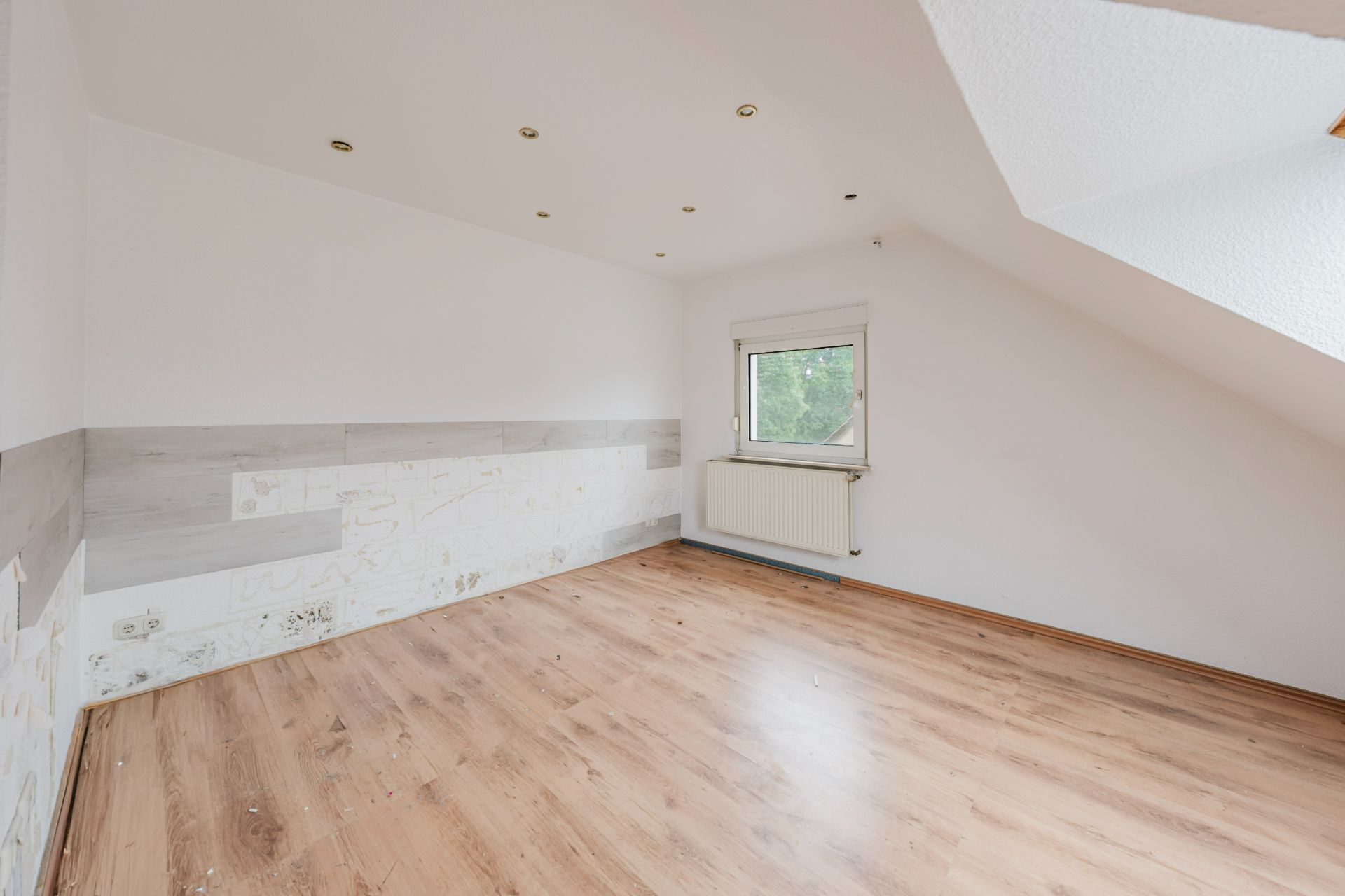 Leeres Zimmer mit Holzfußboden, schräger Decke und kleinem Fenster mit Heizkörper darunter.