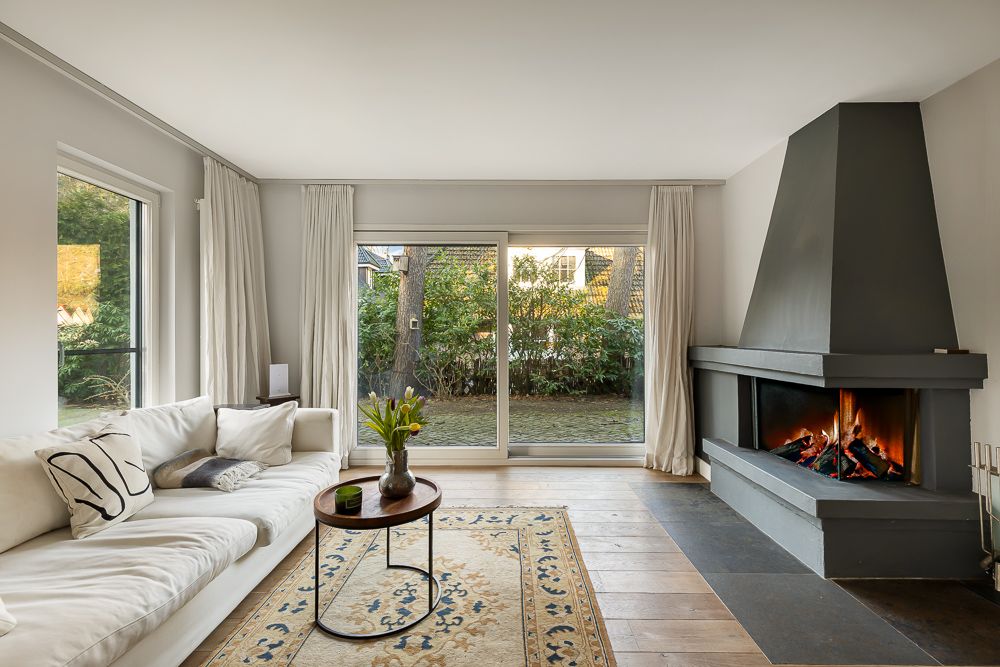 Helles Wohnzimmer mit Sofa, Teppich, Tisch und Kamin, große Fenster mit Blick in den Garten.