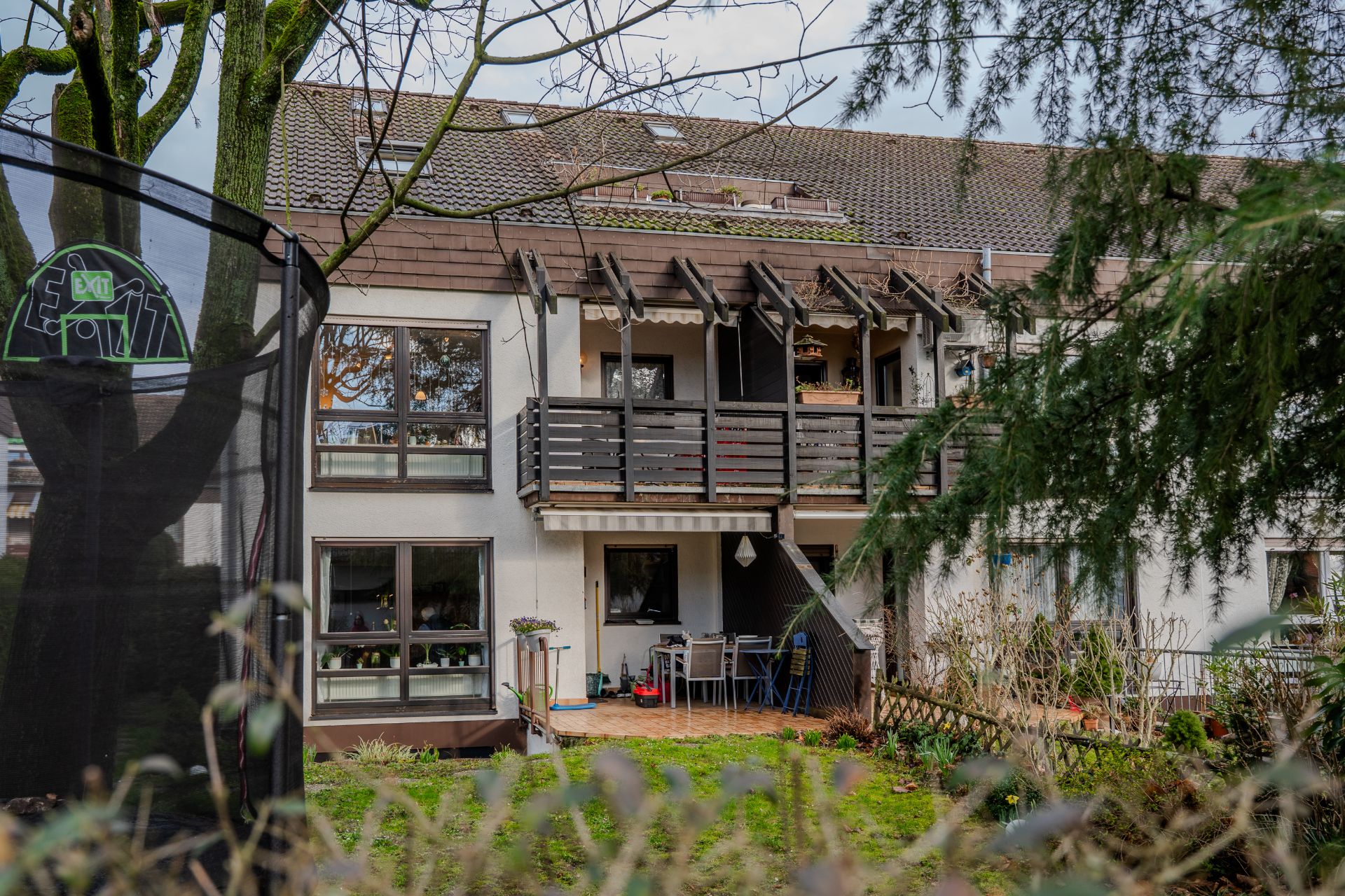 Mehrfamilienhaus mit Balkon, Terrasse und Garten, im Vordergrund ein Trampolin und Bäume.