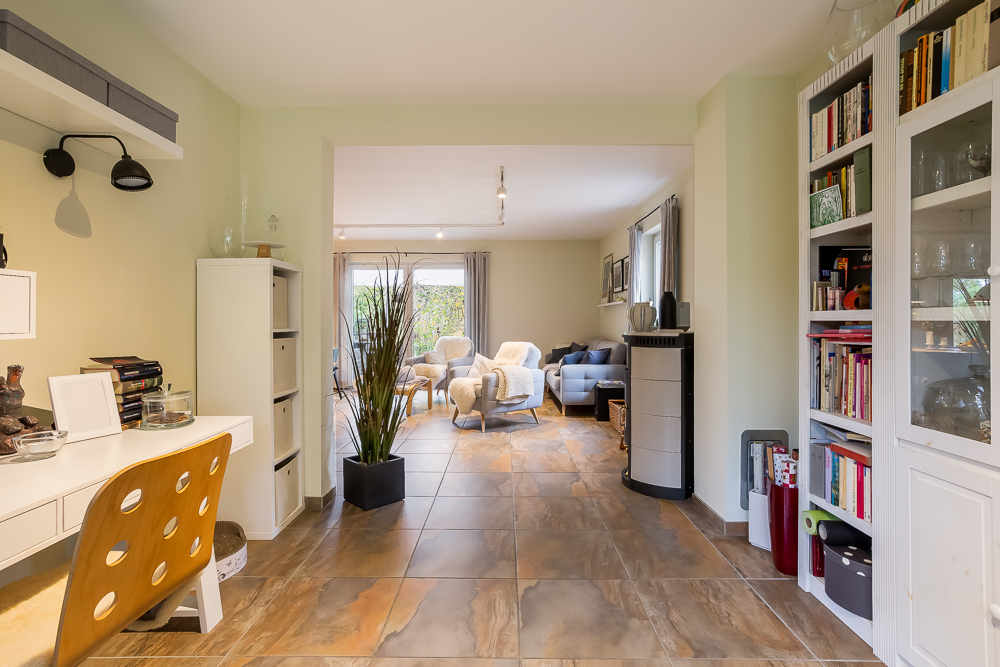 Helles Wohnzimmer mit Schreibtisch, Bücherregalen, Sesseln und großer Fensterfront.