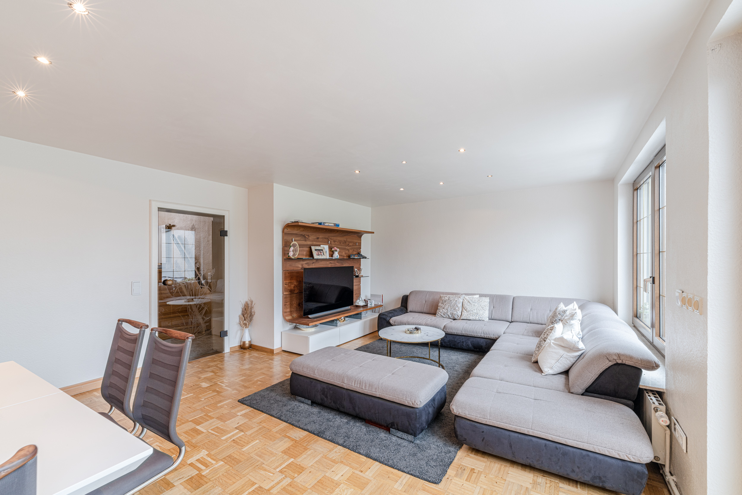 Helles Wohnzimmer mit großer Eckcouch, Glastisch, TV und Holzregal auf Parkettboden.
