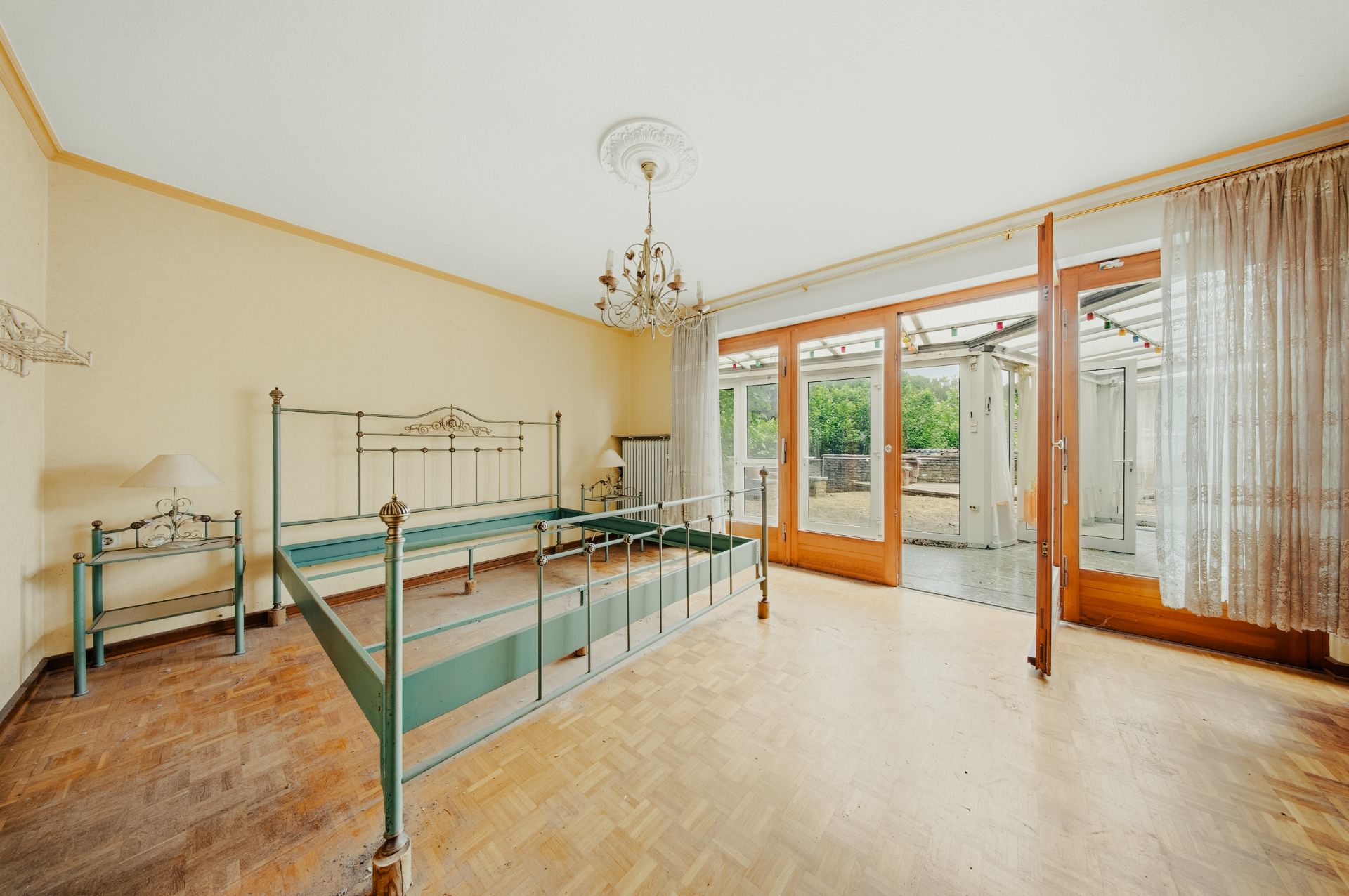 Helles Schlafzimmer mit Metallbettgestell, Nachttisch, Kronleuchter und großen Fenstern zur Terrasse.