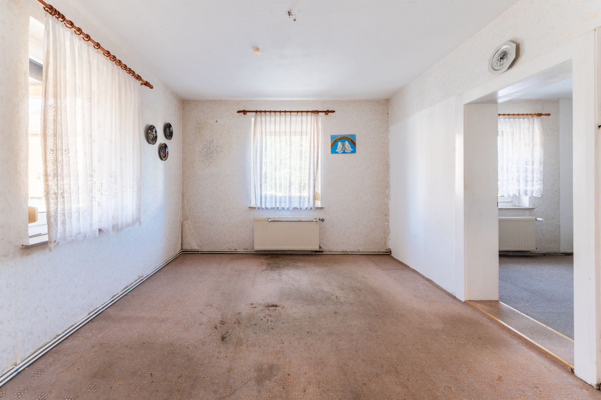 Leeres Wohnzimmer mit zwei Fenstern, Vorhängen und Teppichboden, Zugang zu angrenzendem Raum sichtbar.