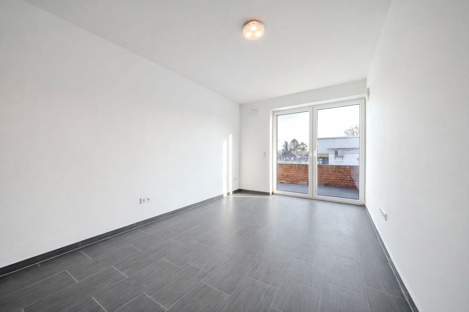 Helles, leeres Zimmer mit grauem Fliesenboden und großen Fenstertüren zum Balkon.