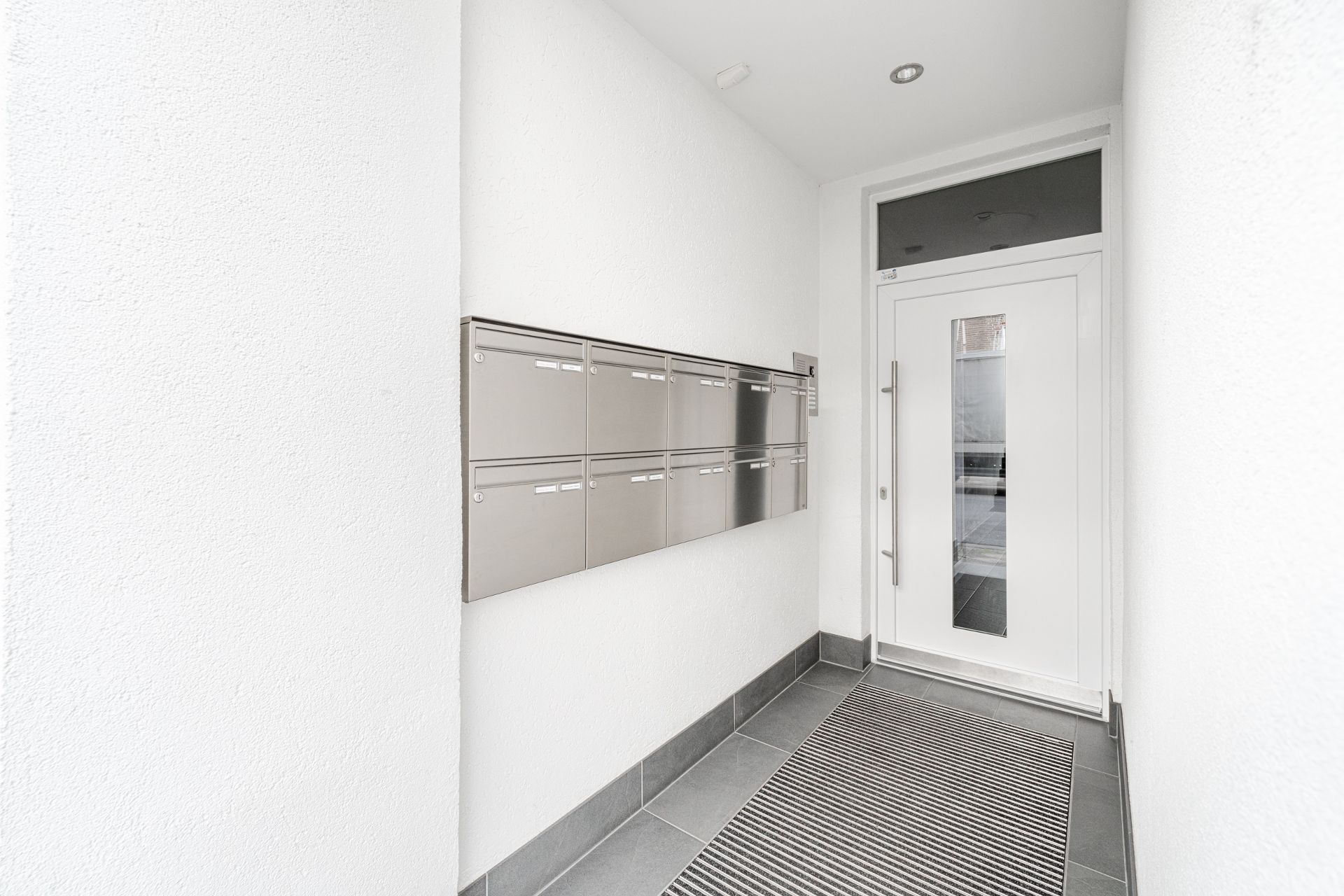 Weiße Hauseingangstür mit mehreren Briefkästen an der Wand und Fußmatte auf dem Boden.