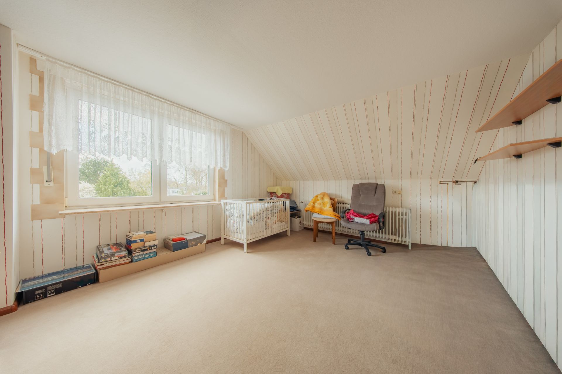 Helles Zimmer mit schräger Decke, Babybett, Sessel, Regalen und großen Fenstern.