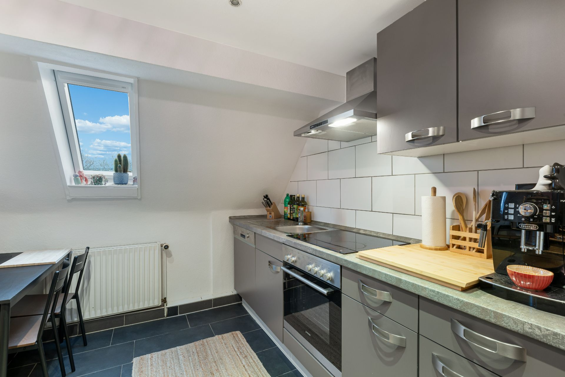 Moderne Küche mit grauen Schränken, Einbauherd, Kaffeemaschine und Esstisch am Fenster.
