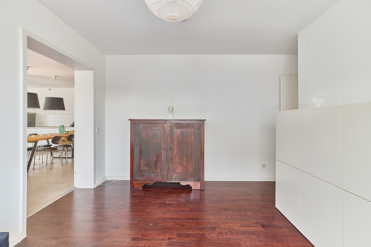 Helles Zimmer mit dunklem Holzschrank, Holzboden und weißer Wand, Blick ins Esszimmer.
