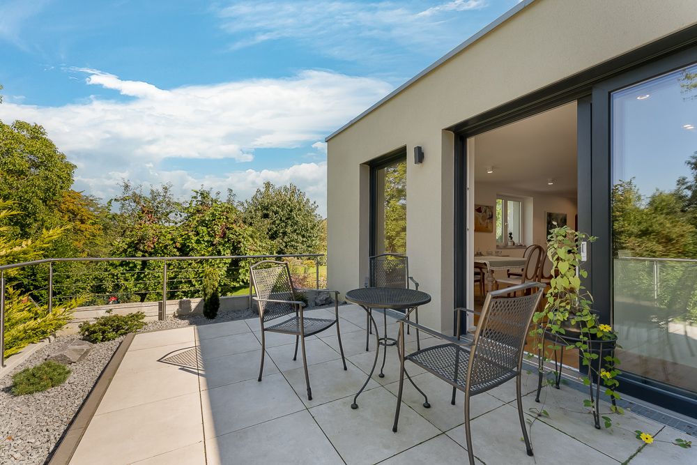 Moderne Terrasse mit Metalltisch und Stühlen, Blick ins Grüne, große Glasschiebetür zum Wohnbereich.