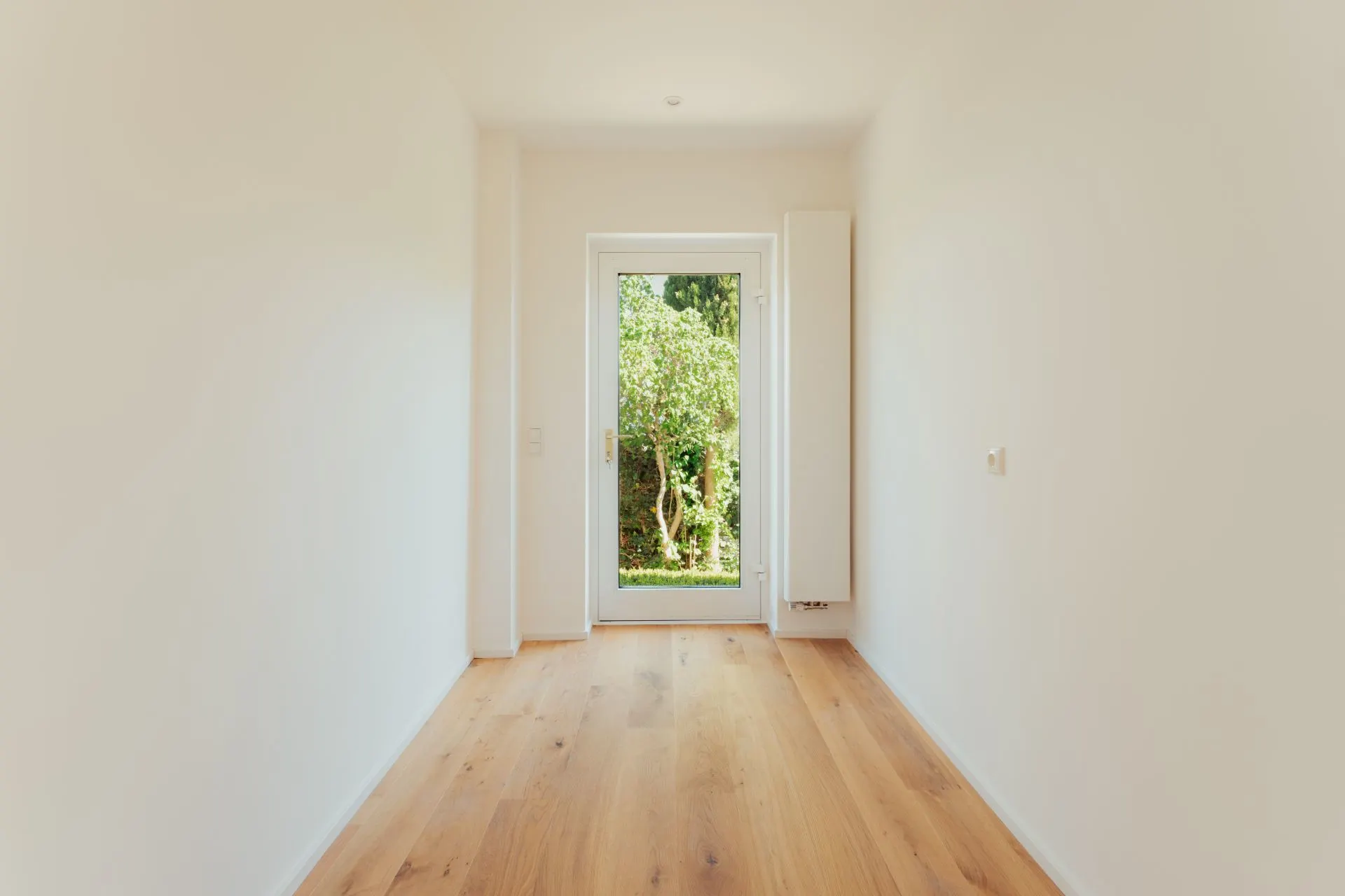 Heller, leerer Raum mit Holzfußboden und Glastür zu einem Garten.