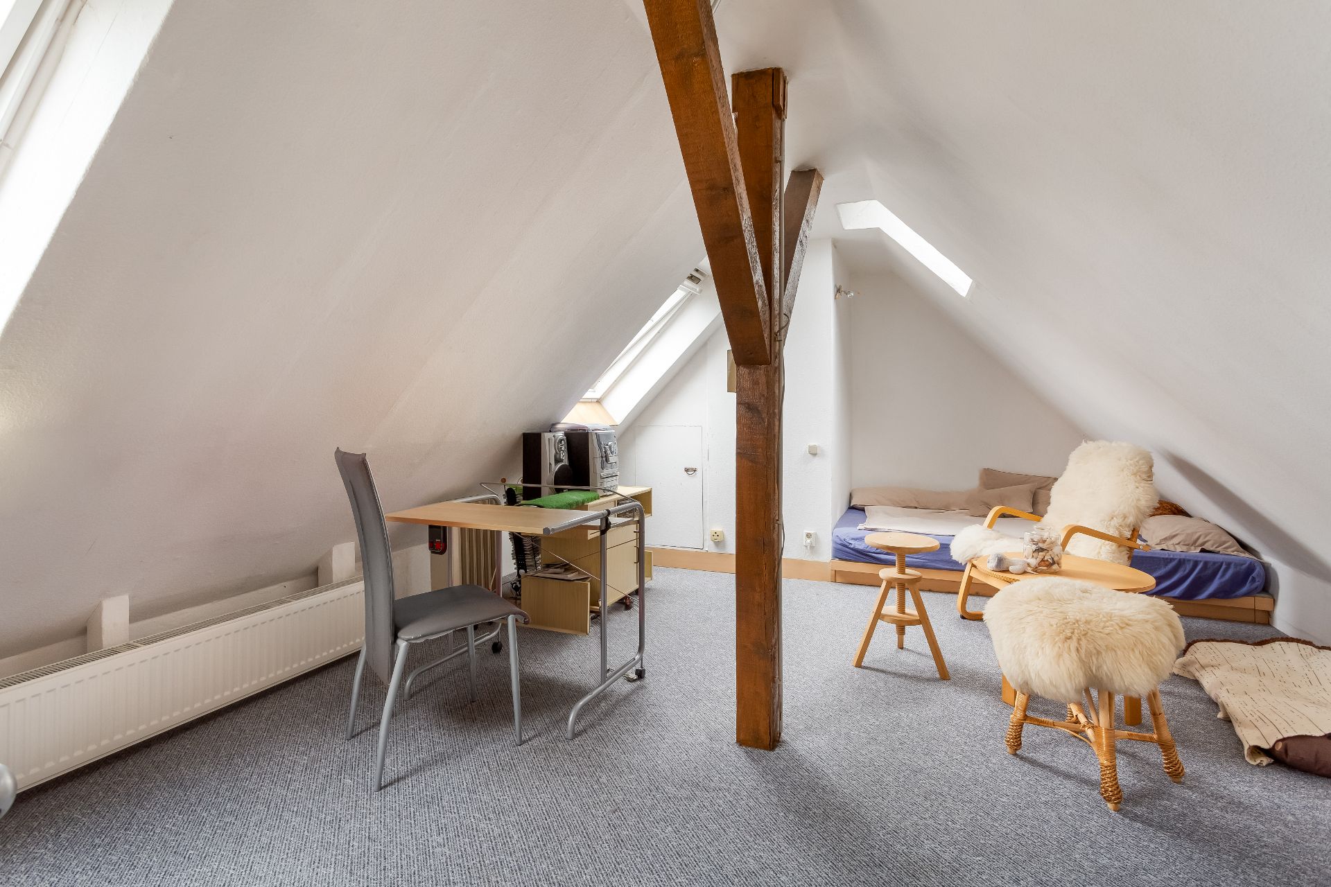 Dachzimmer mit Schreibtisch, Stuhl, Bett und Sessel mit Fellauflage, sichtbarem Holzbalken.