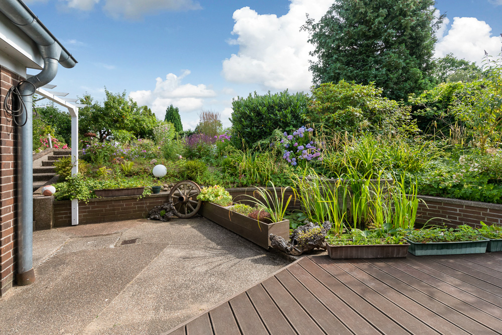 Terrasse mit Hochbeeten, Blumen, Sträuchern und altem Wagenrad im Garten.