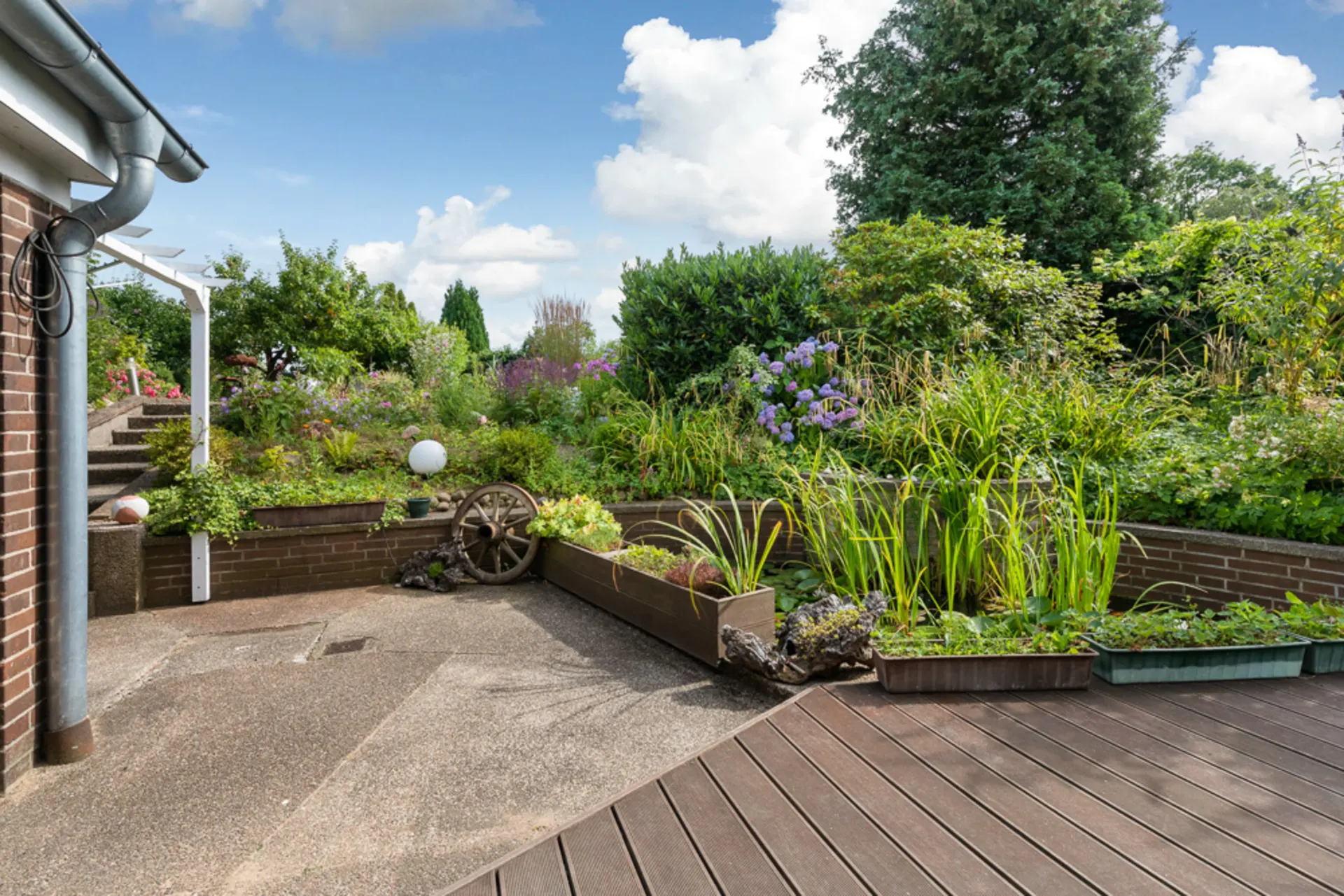 Terrasse mit Hochbeeten, Blumen, Sträuchern und altem Wagenrad im Garten.
