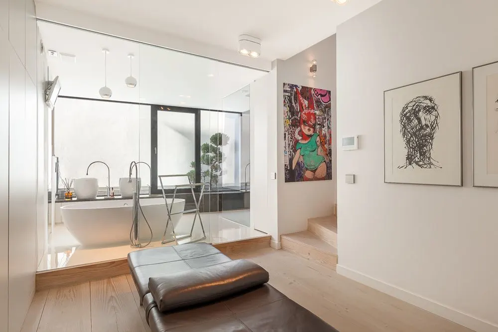 Modernes Badezimmer mit freistehender Badewanne hinter Glaswand, Loungemöbel und bunter Wandkunst.