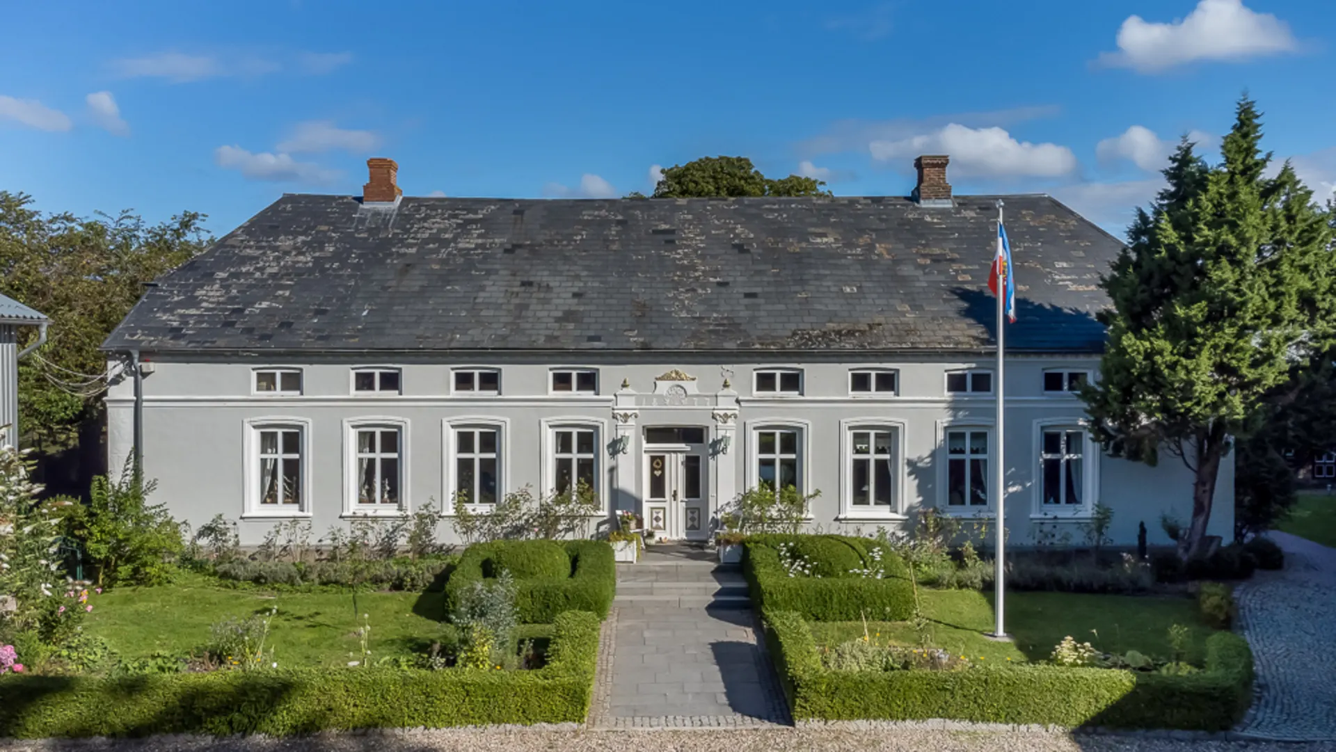 Großes weißes Wohnhaus mit Garten, Büschen, Gehweg und Fahnenmast an der Vorderseite.