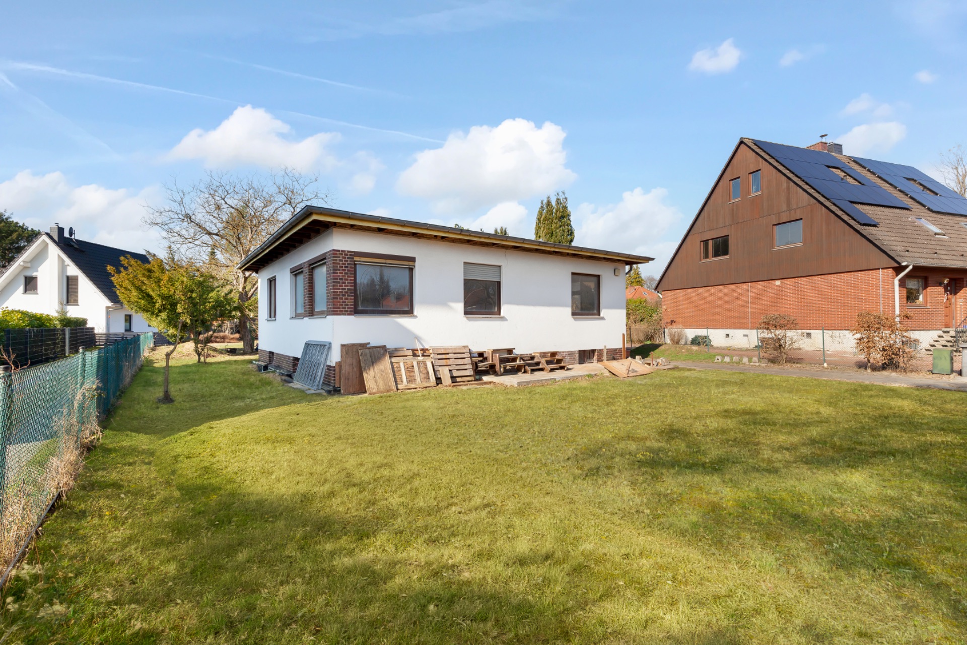 Einfache weiße Flachdachhaus mit großem Garten und Zaun, daneben ein weiteres Wohnhaus.