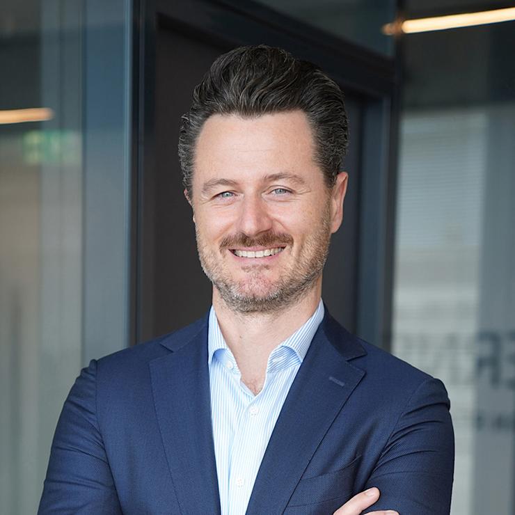 Mann im Anzug steht mit verschränkten Armen in einem modernen Innenraum.