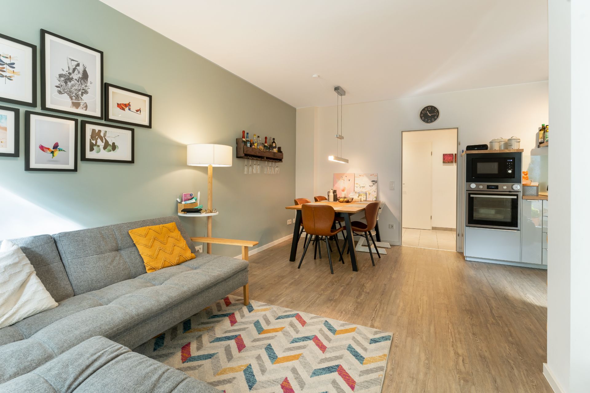 Helles Wohnzimmer mit grauem Sofa, Esstisch, offener Küche und Bilderwand.