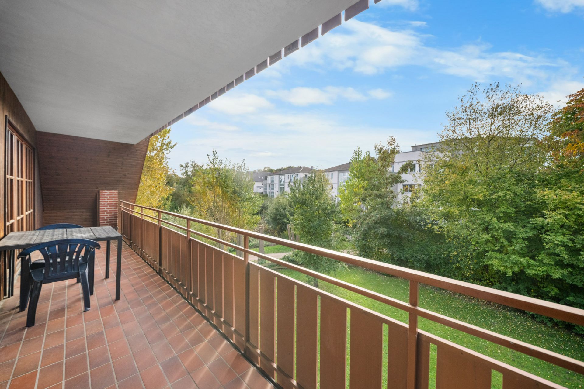 Großer Balkon mit Tisch und Stuhl, Blick auf grüne Bäume und Wohngebäude im Hintergrund.