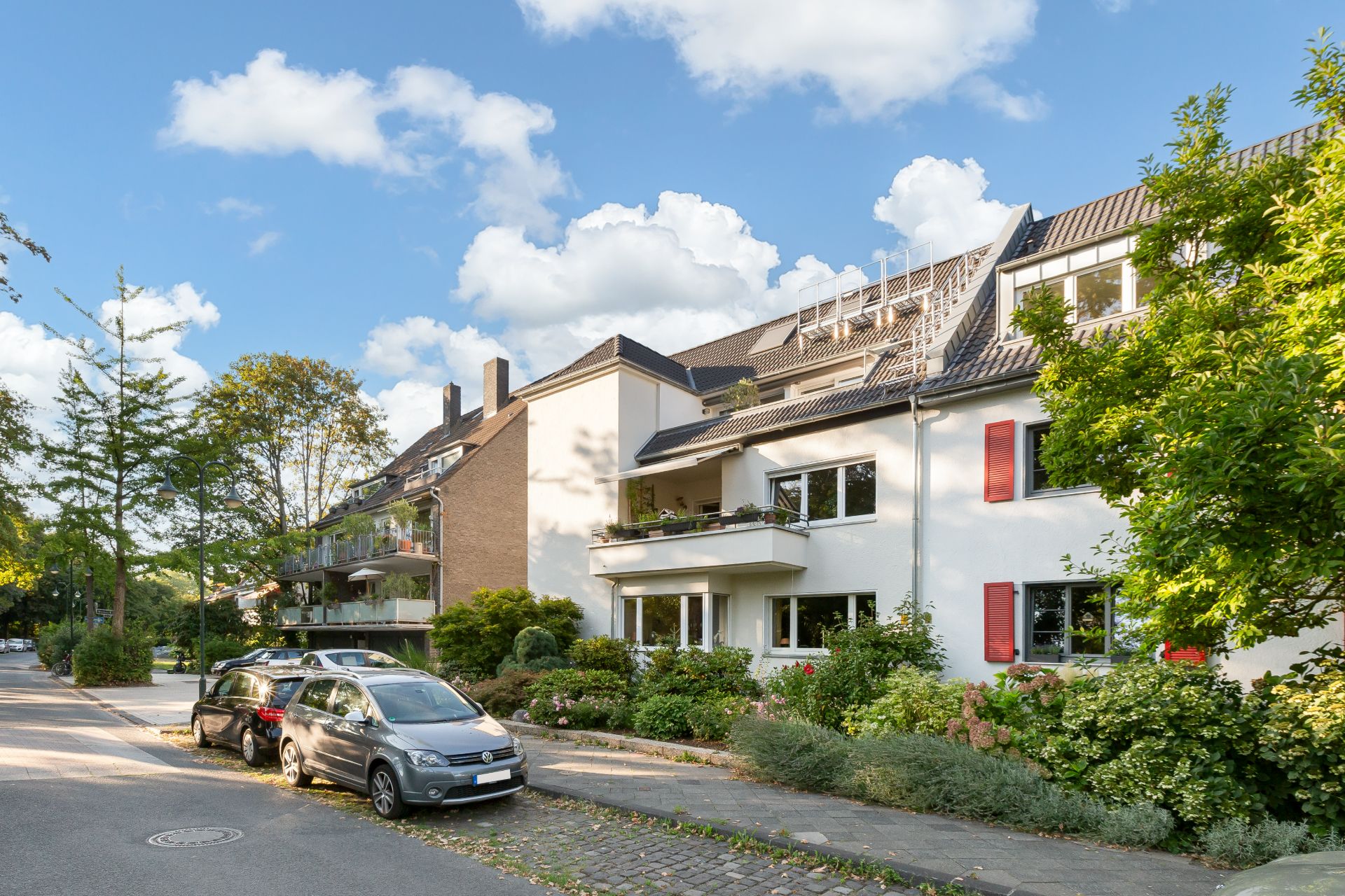 Mehrfamilienhaus mit Balkonen und Vorgarten an einer ruhigen Wohnstraße, Autos parken am Straßenrand.