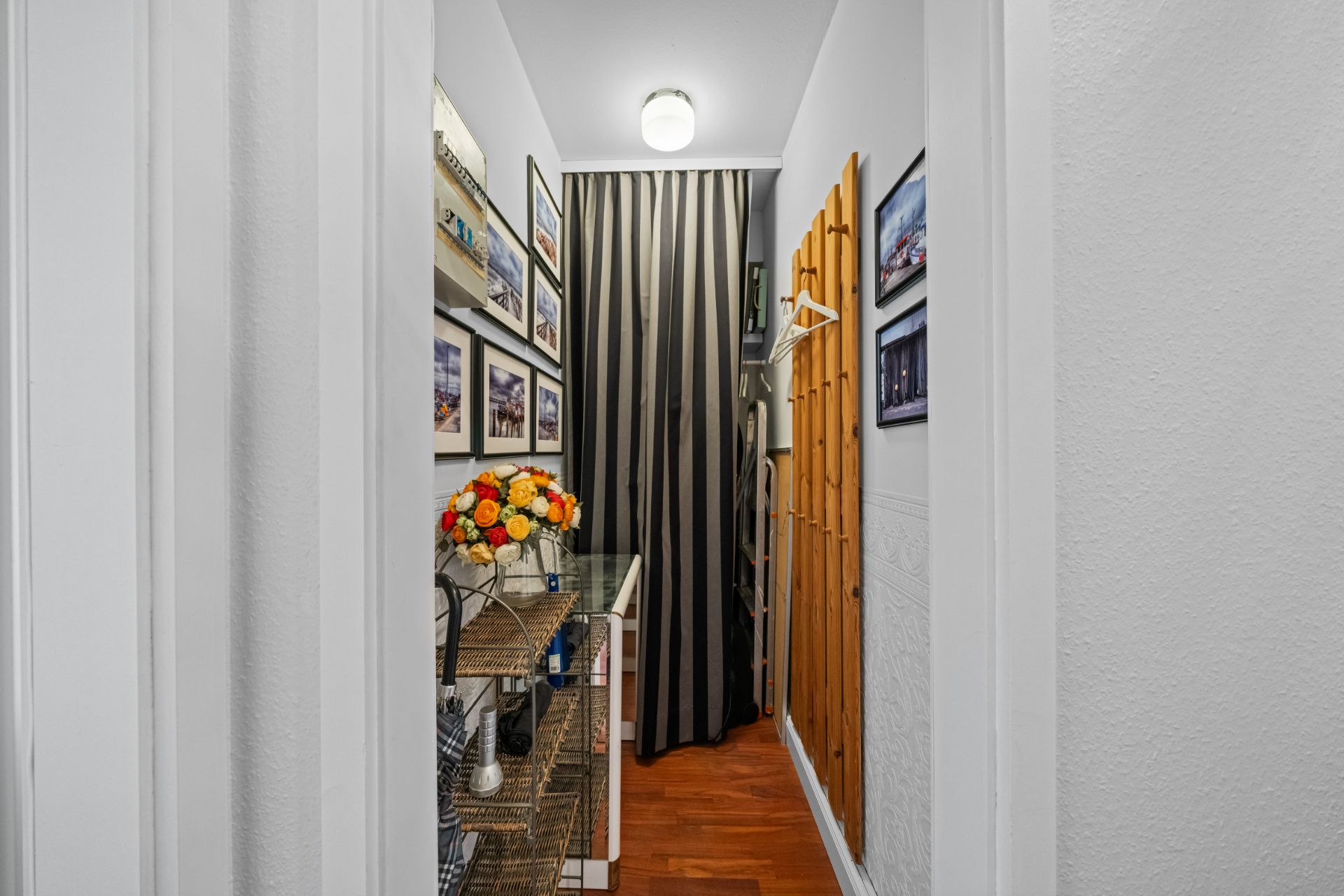 Schmaler Flur mit Wandbildern, Regal, Blumenstrauß und Garderobe auf Holzboden.