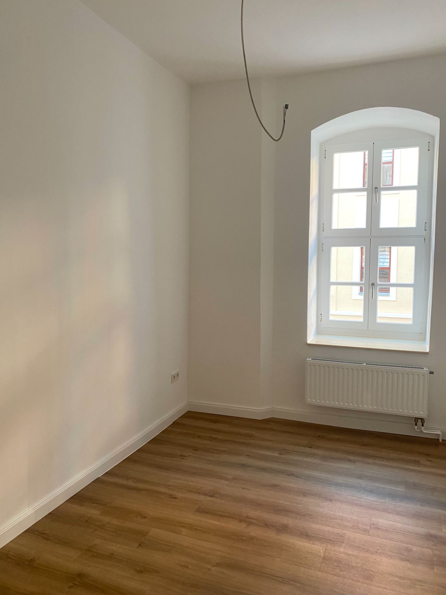 Leeres Zimmer mit Holzboden, weißer Wand, Fenster und Heizkörper.