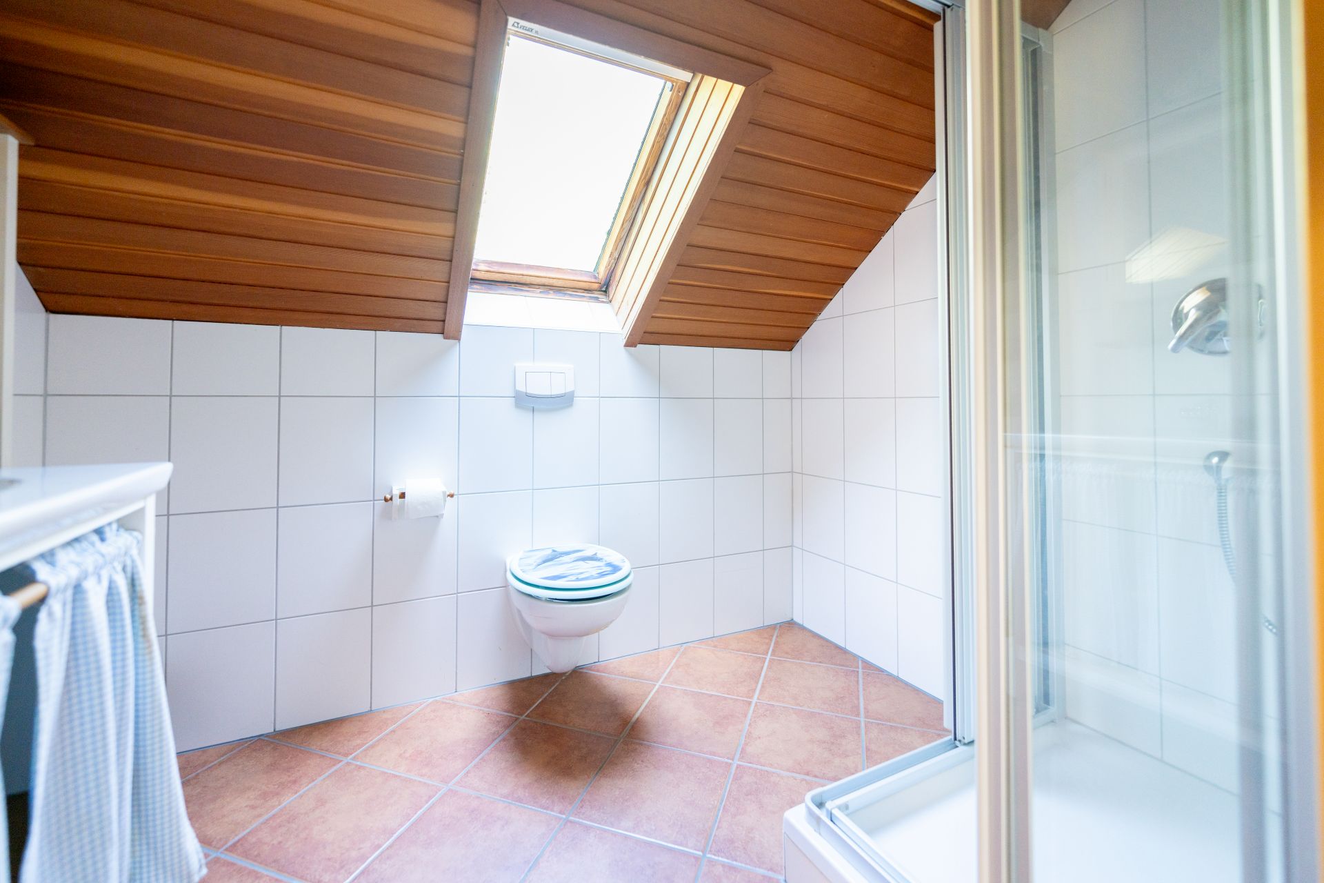 Helles Bad mit Dachfenster, Duschkabine und wandhängender Toilette auf Fliesenboden.