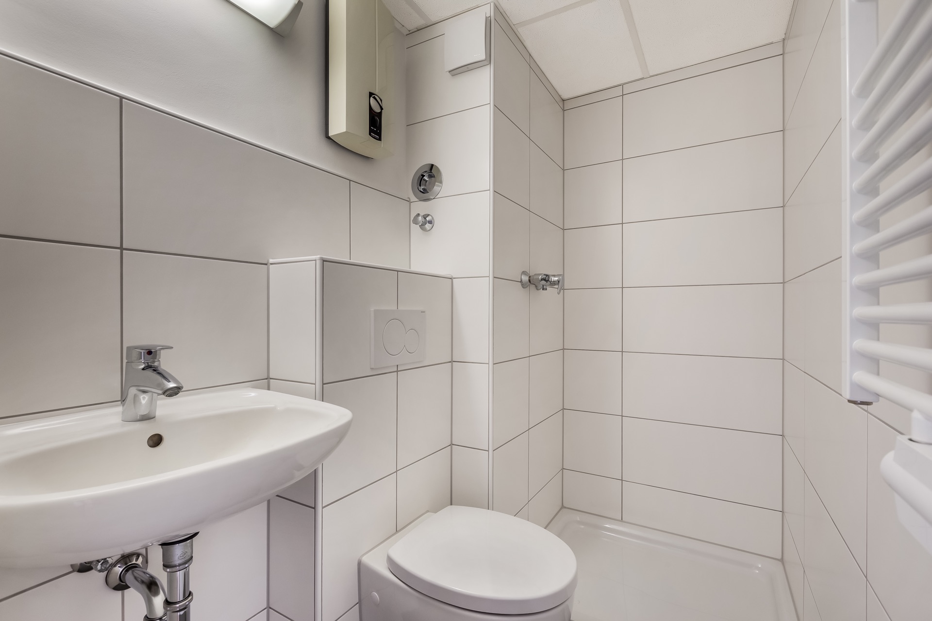 Kleines Badezimmer mit Waschbecken, Toilette und Dusche, alles in weißen Fliesen gehalten.