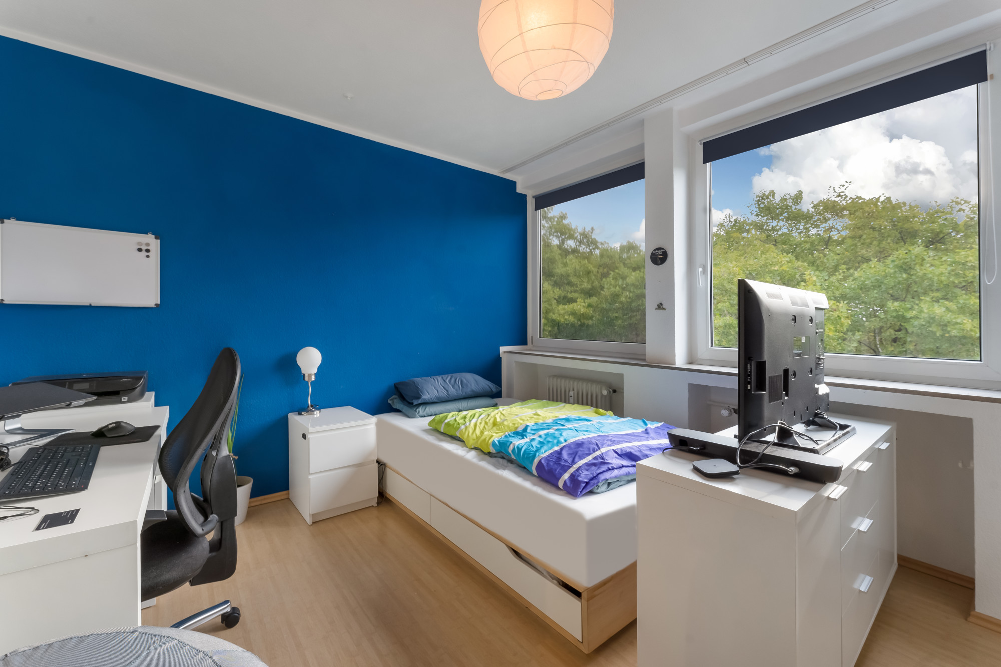 Helles Schlafzimmer mit Schreibtisch, Einzelbett, Kommode und großem Fenster mit Blick auf Bäume.