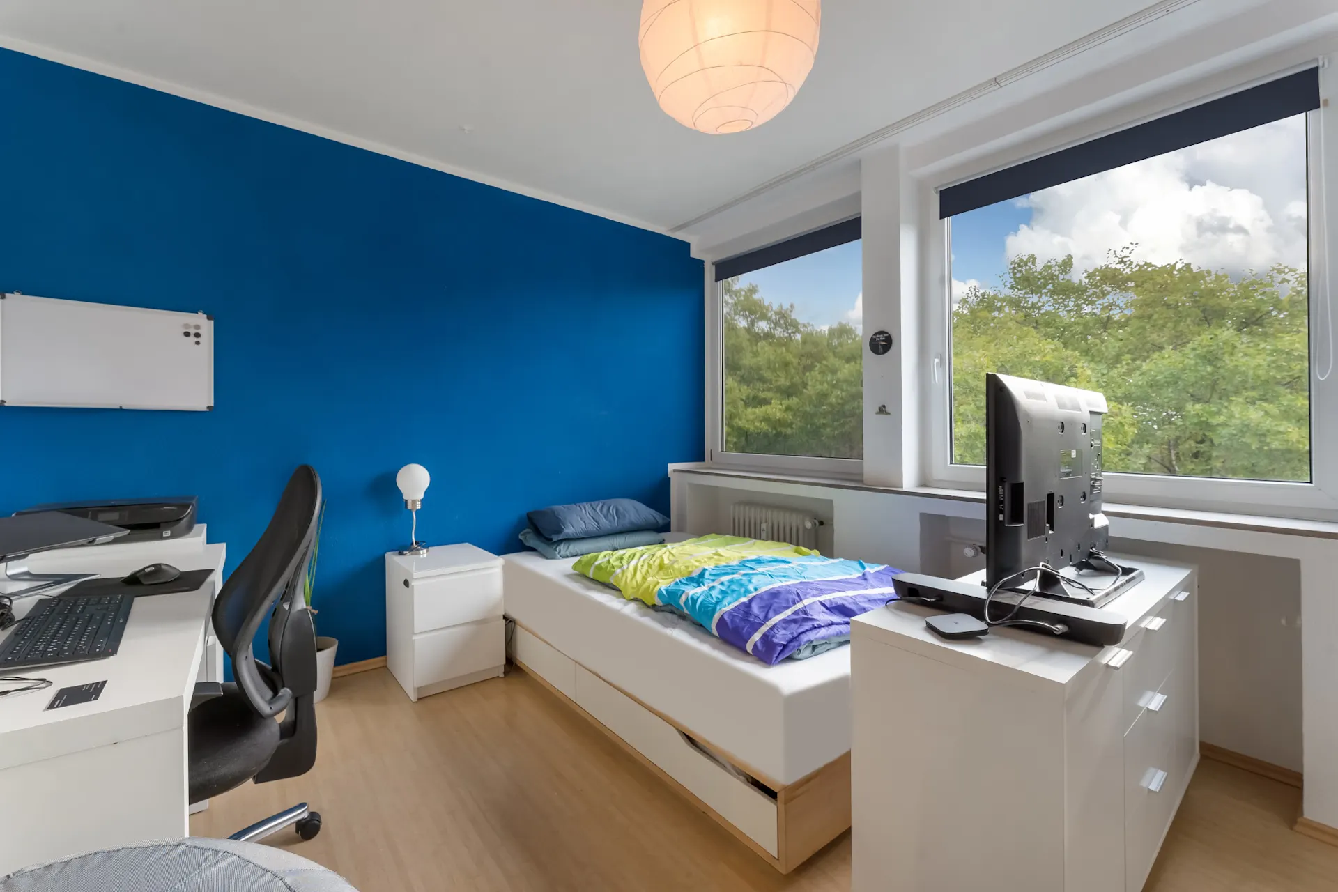 Helles Schlafzimmer mit Schreibtisch, Einzelbett, Kommode und großem Fenster mit Blick auf Bäume.