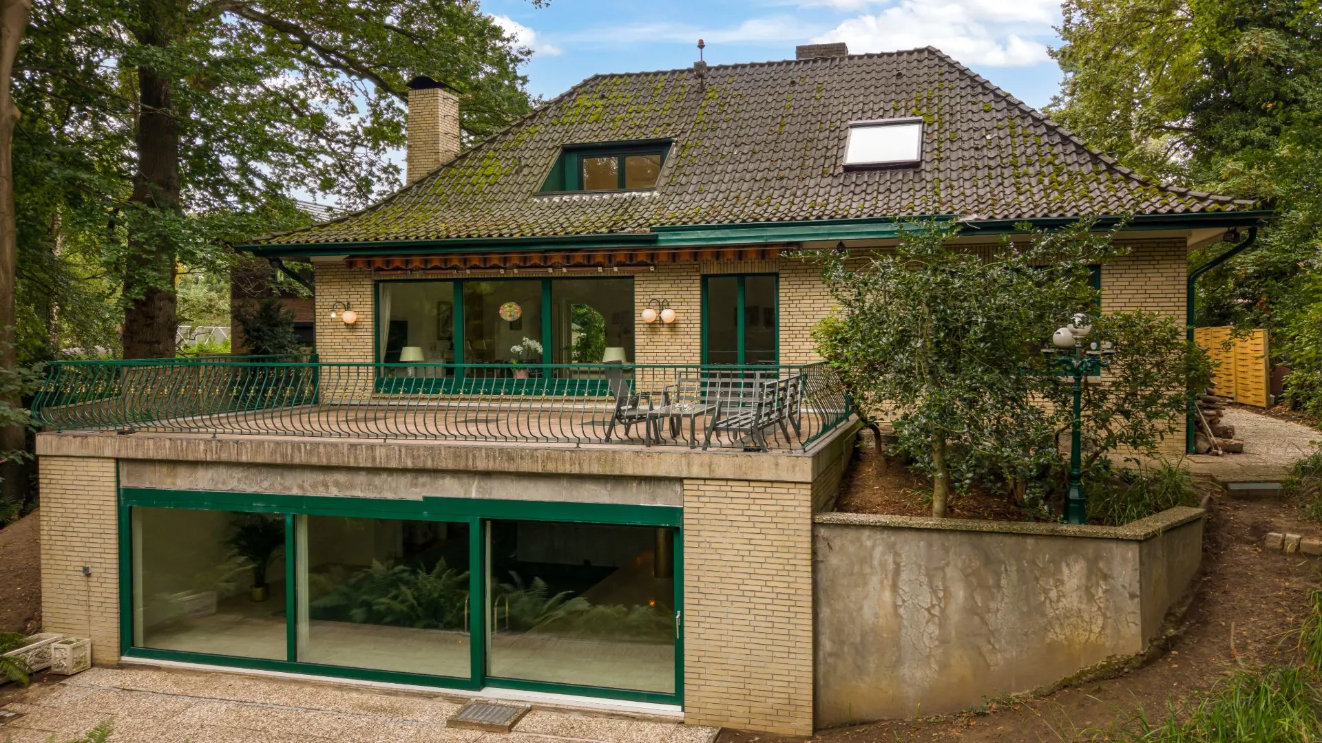 Zweistöckiges Haus mit großer Terrasse, grünem Geländer und vielen Fenstern, umgeben von Bäumen.