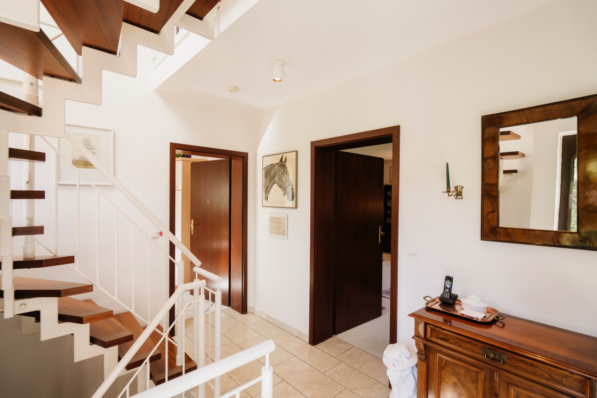 Heller Flur mit Holztreppe, zwei offenen Türen, Kommode und Spiegel an der Wand.