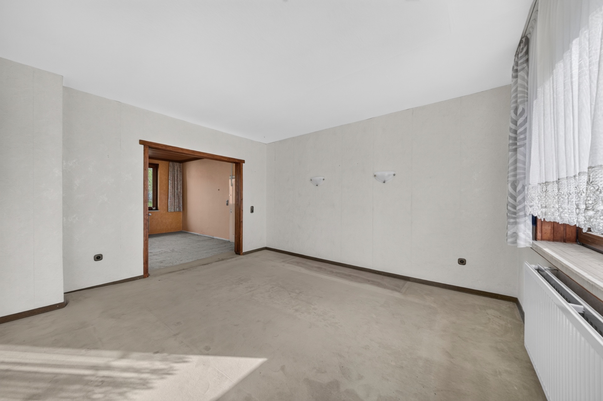 Leeres Zimmer mit beigem Teppich, großem Fenster und Durchgang zum nächsten Raum.