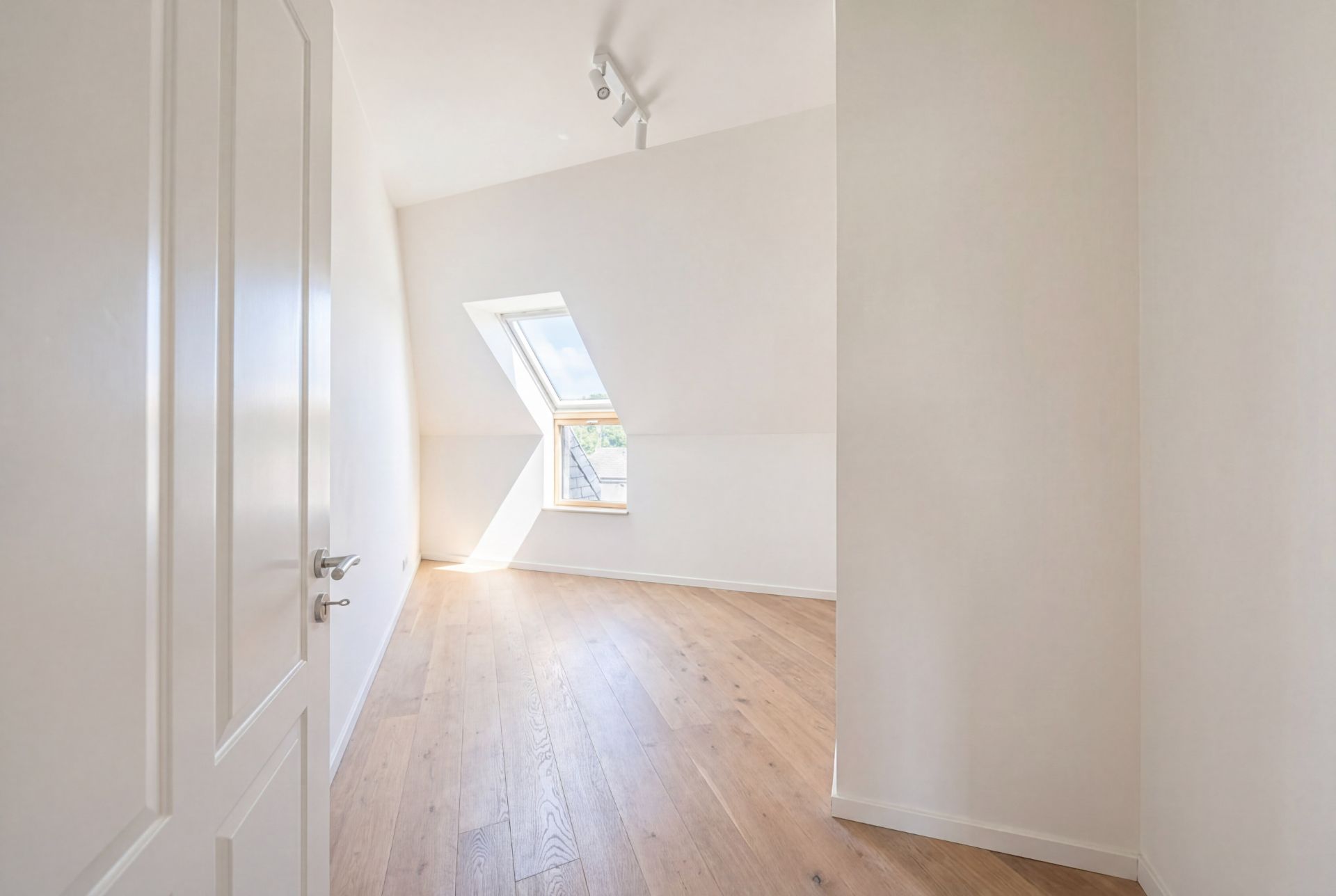 Heller Raum mit weißer Tür, Schräge, großem Fenster und Holzfußboden.