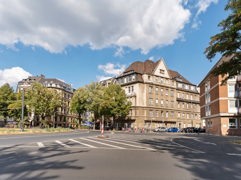 Großes Wohngebäude an einer Kreuzung mit Bäumen und geparkten Autos.
