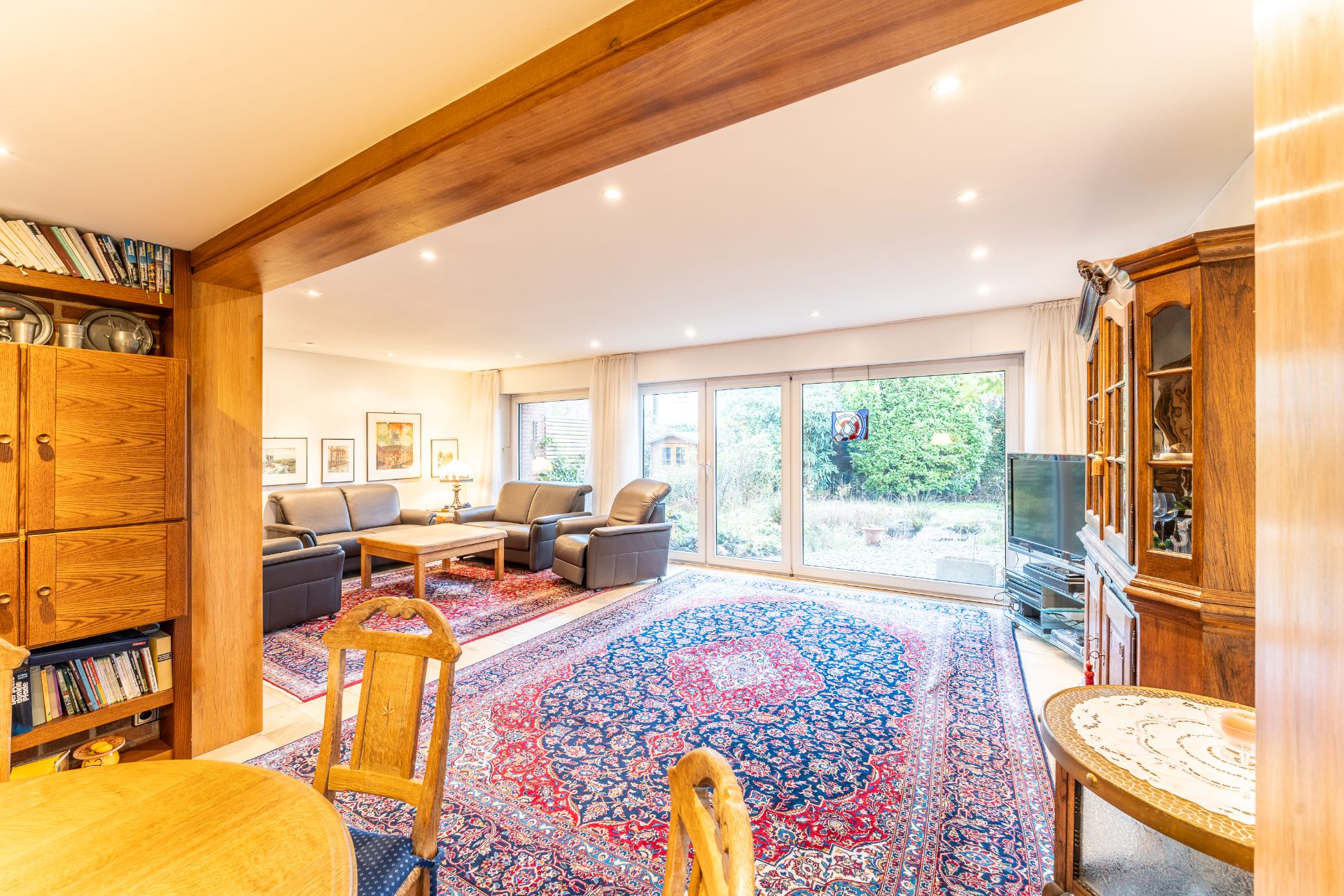 Helles Wohnzimmer mit großen Fenstern, Teppichen, Ledersofas, Holzmöbeln und Blick in den Garten.